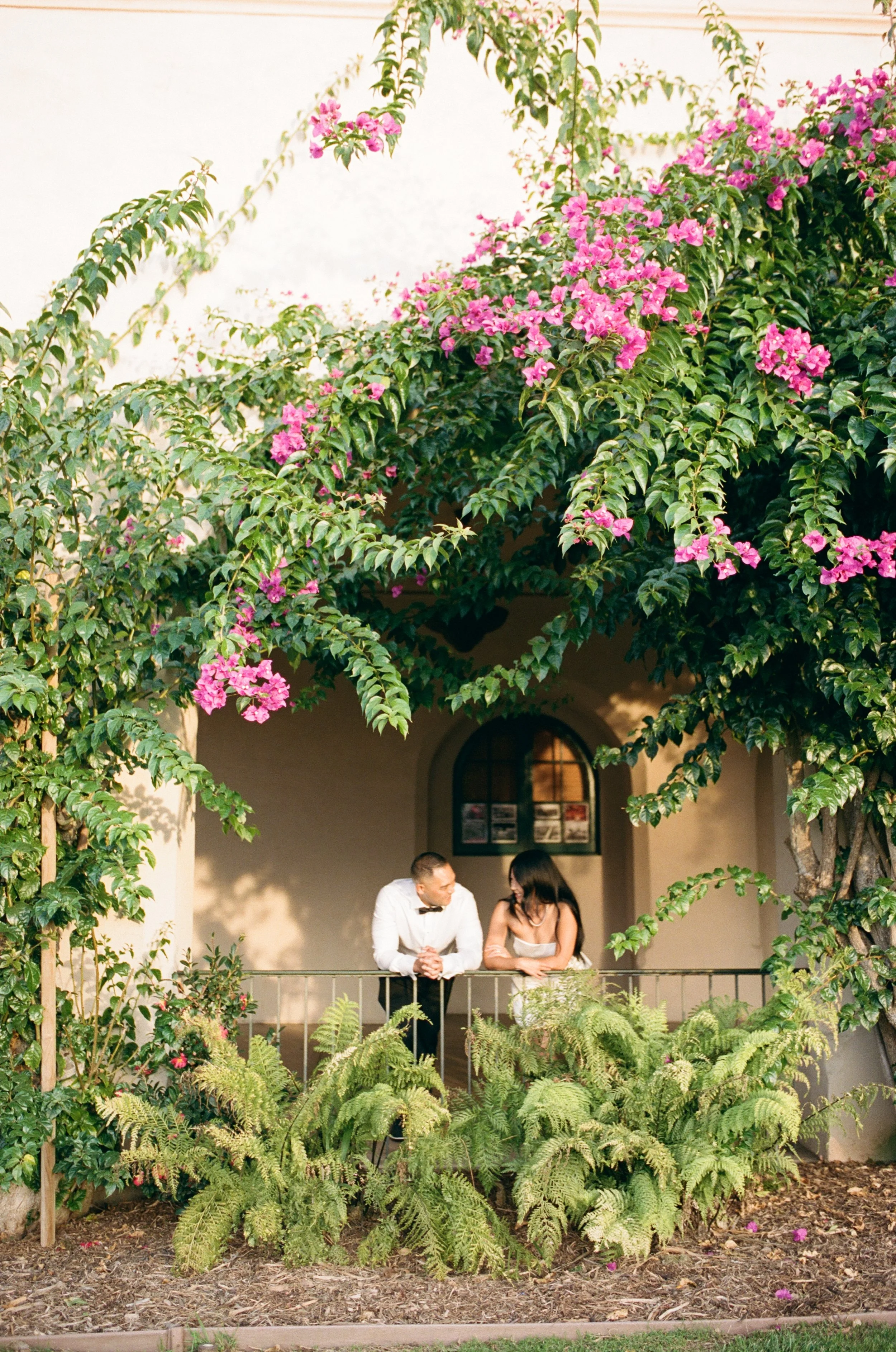 A man in a tuxedo and a woman in a white dress with black trim talking on a balcony surrounded by greenery and pink flowering plants.