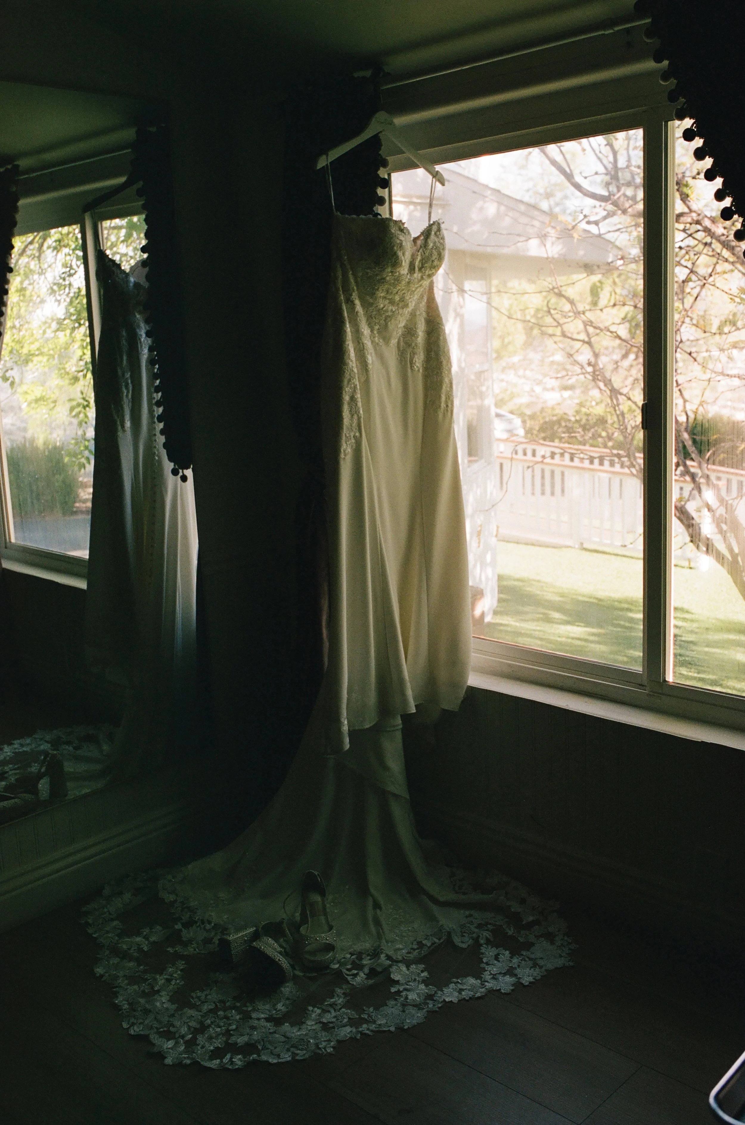 A wedding dress hanging on a hanger near a large window, with sunlight illuminating the dress and a garden with trees visible outside.