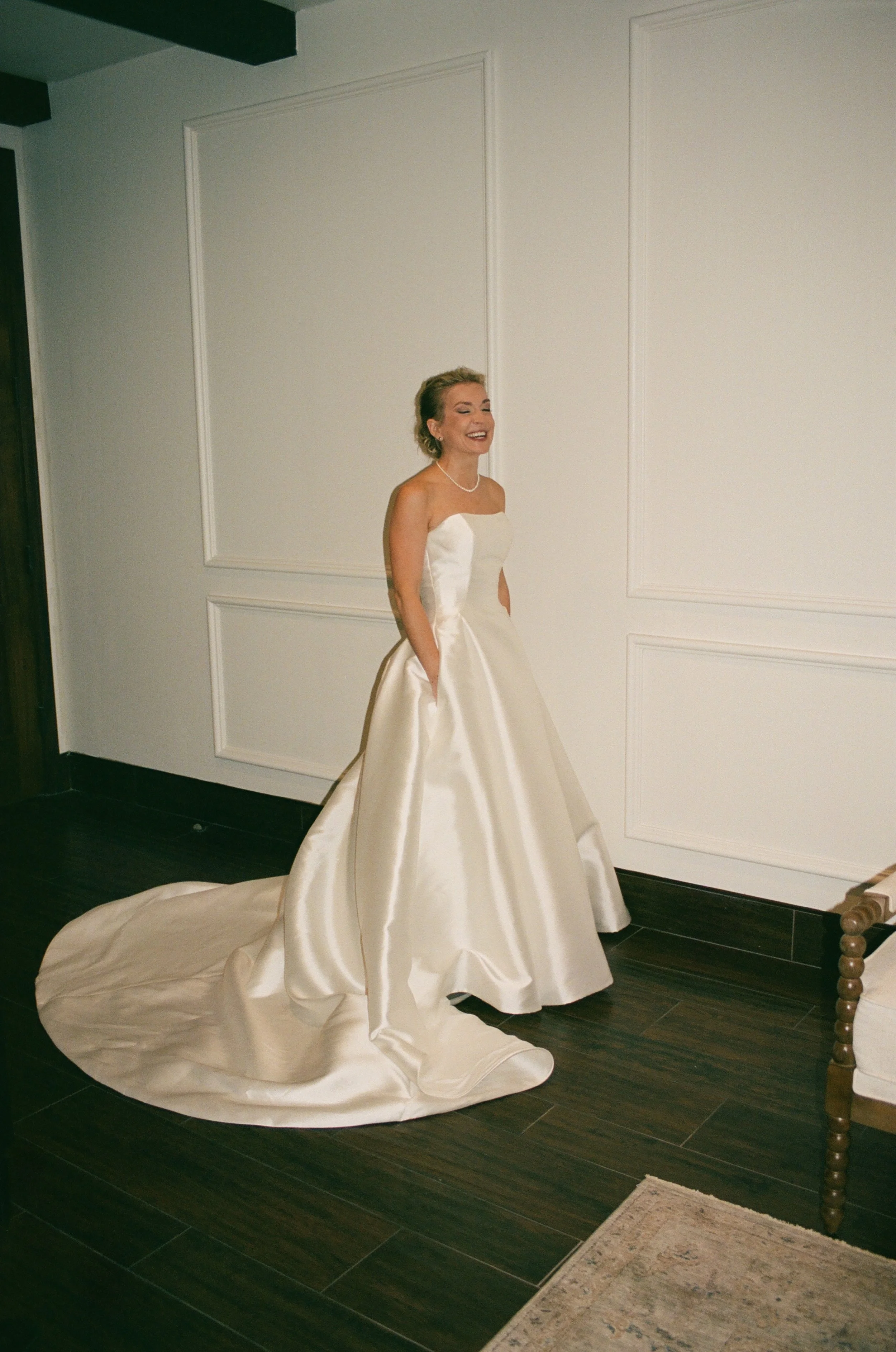 A woman in a strapless white wedding gown with a long train, standing indoors against a white paneled wall, smiling and laughing with hands in pockets.