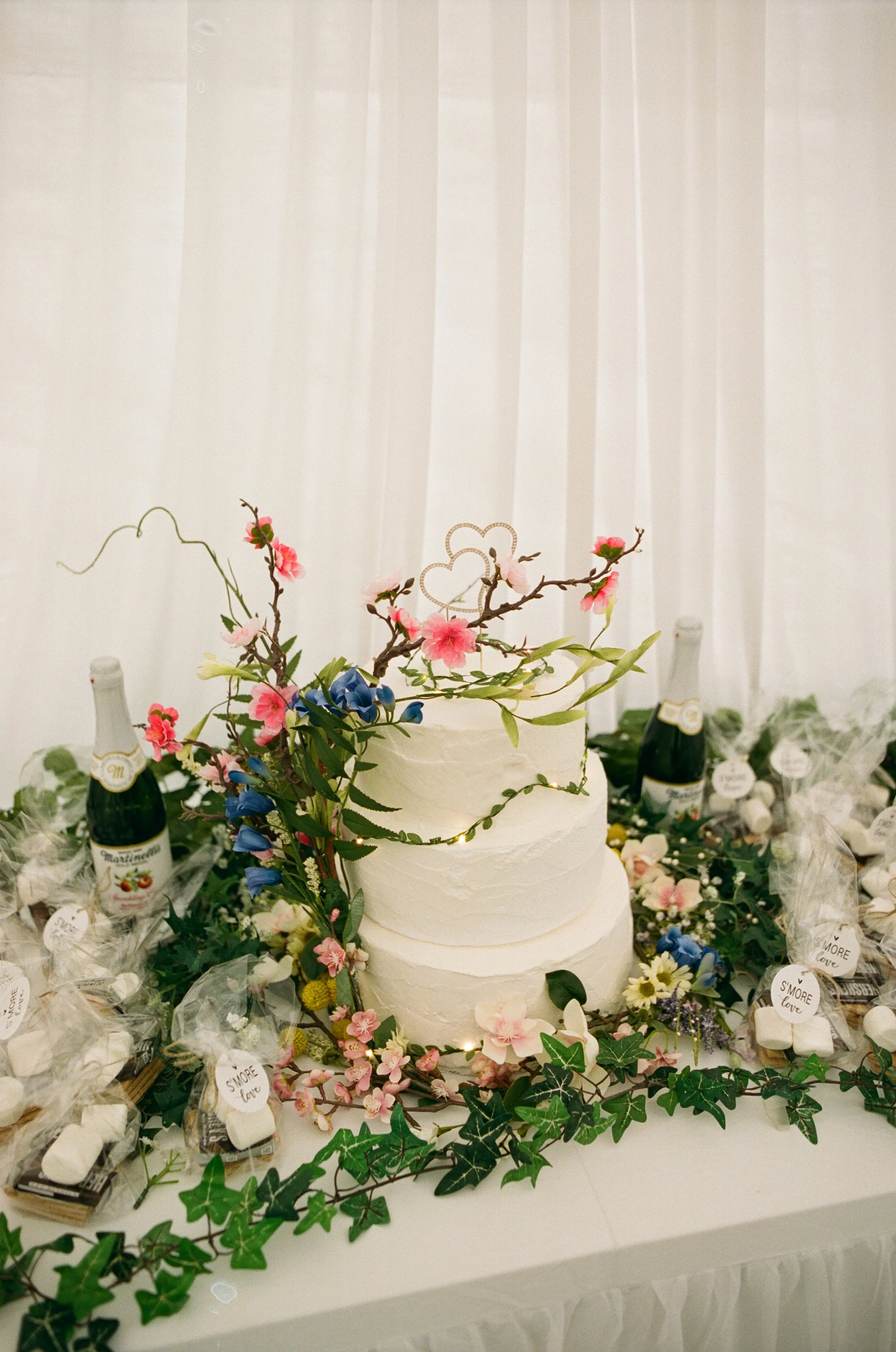Three-tier white wedding cake decorated with pink and blue flowers and green foliage, surrounded by bottles of sparkling wine and wrapped candy on a table with a white tablecloth.