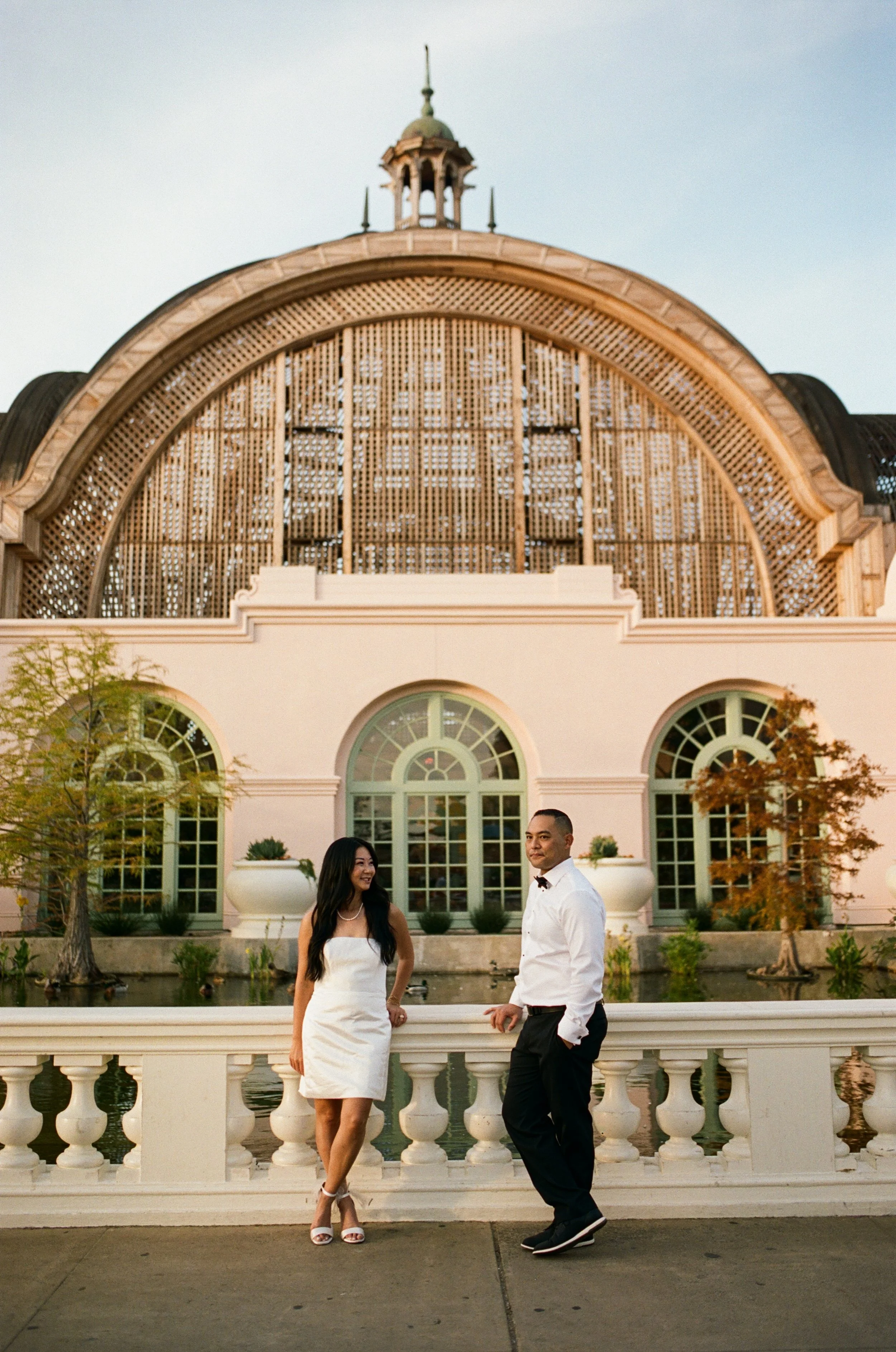 A woman and a man dressed formally, standing on a bridge in front of a building with large arched windows and a decorative wooden facade, during sunset.