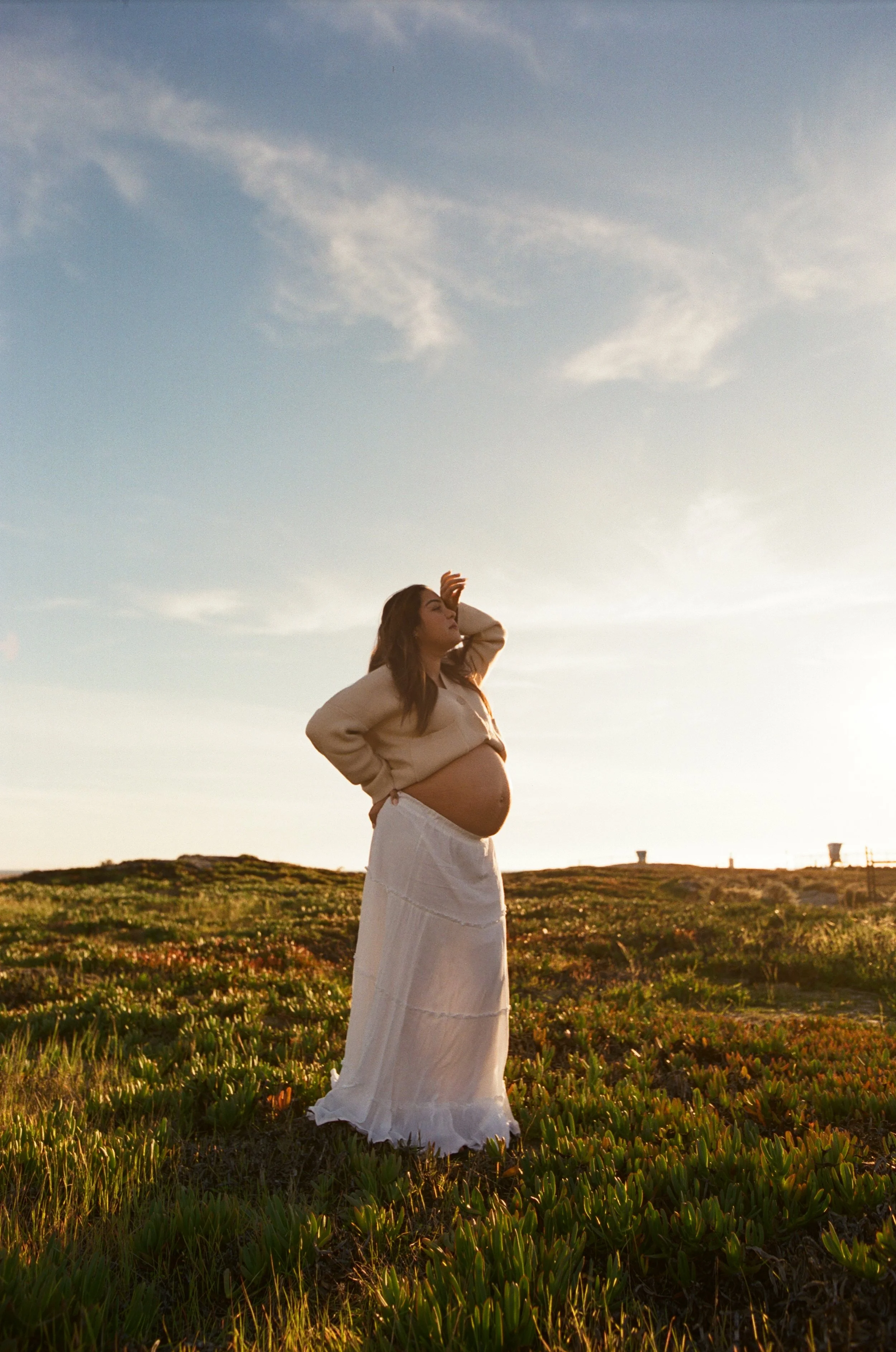 Pregnant woman standing outdoors on a grassy field during sunset, wearing a white skirt and beige top.