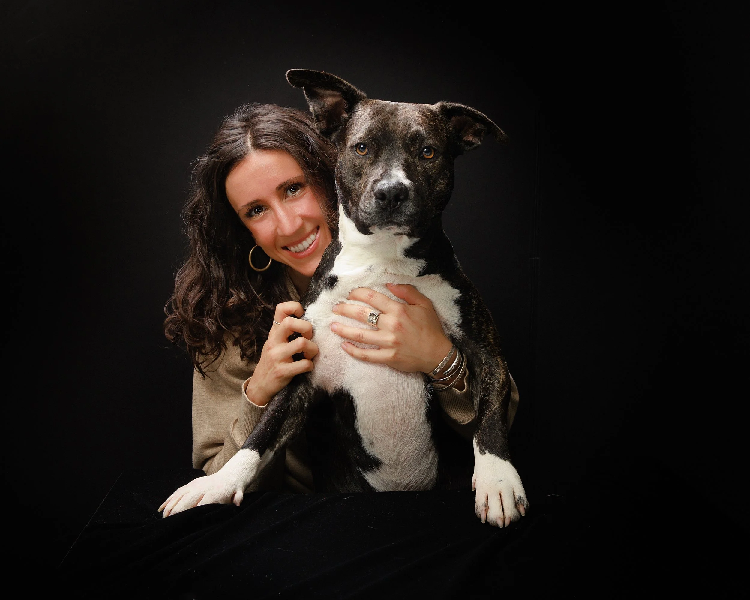 A woman with curly brown hair smiling and hugging a large black and white dog against a black background.