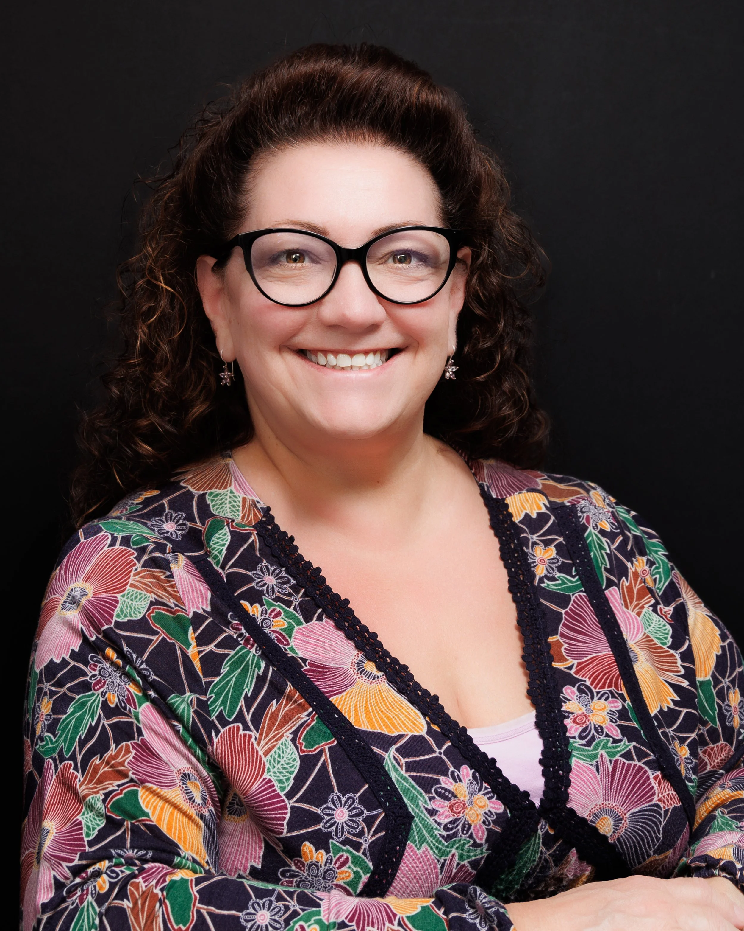 A woman with curly brown hair wearing glasses, a colorful floral patterned top, silver earrings, and smiling at the camera against a black background.