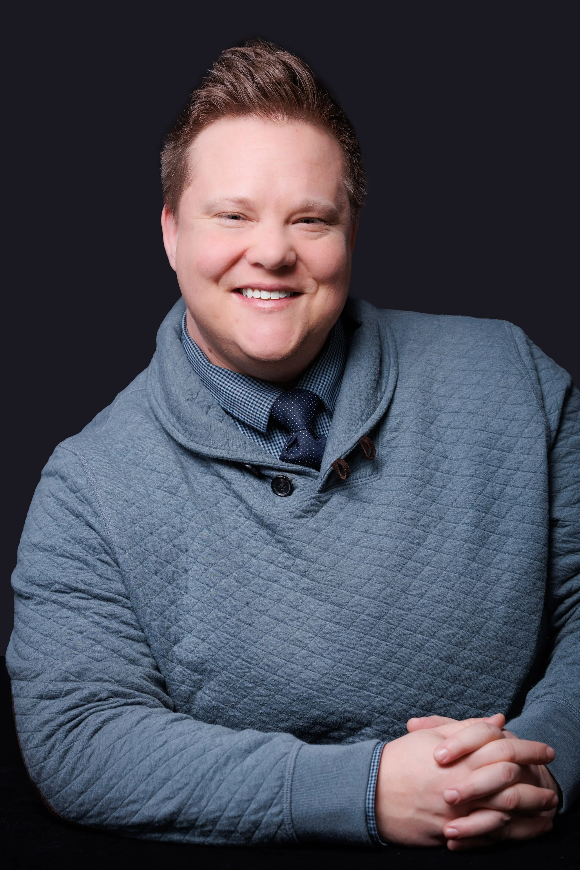 A smiling man with short, styled hair, wearing a blue quilted sweater over a blue checkered shirt and a dark blue tie, posing with folded hands against a dark background.