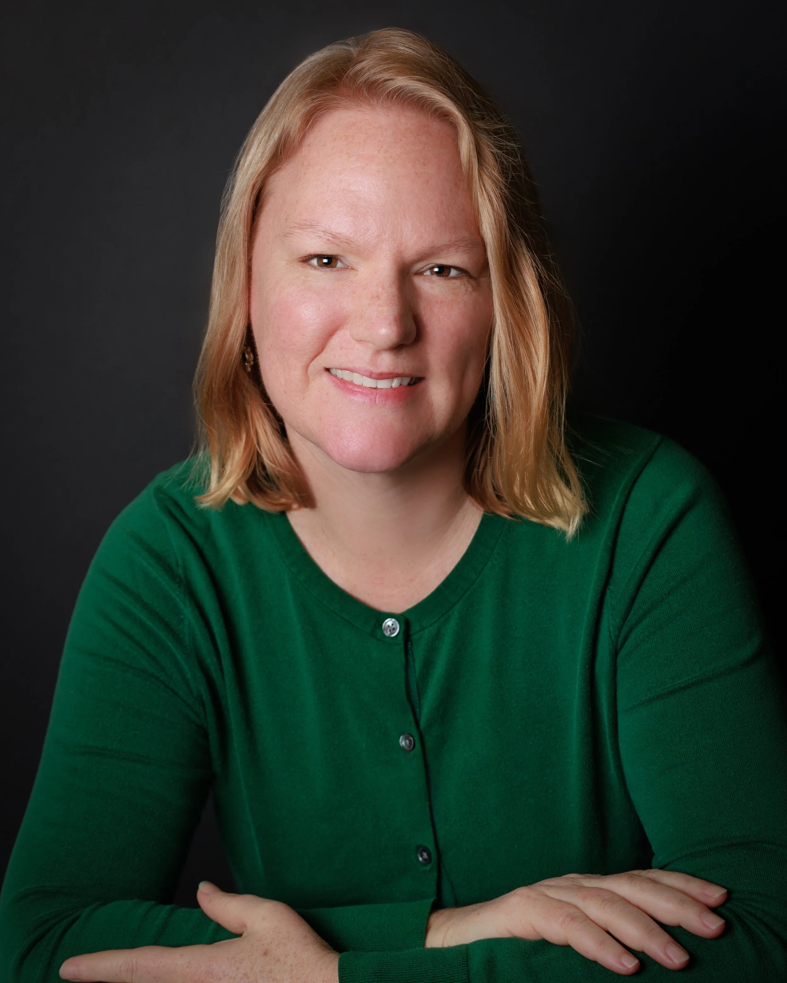 A woman with shoulder-length red hair wearing a green shirt, smiling at the camera, against a dark background.