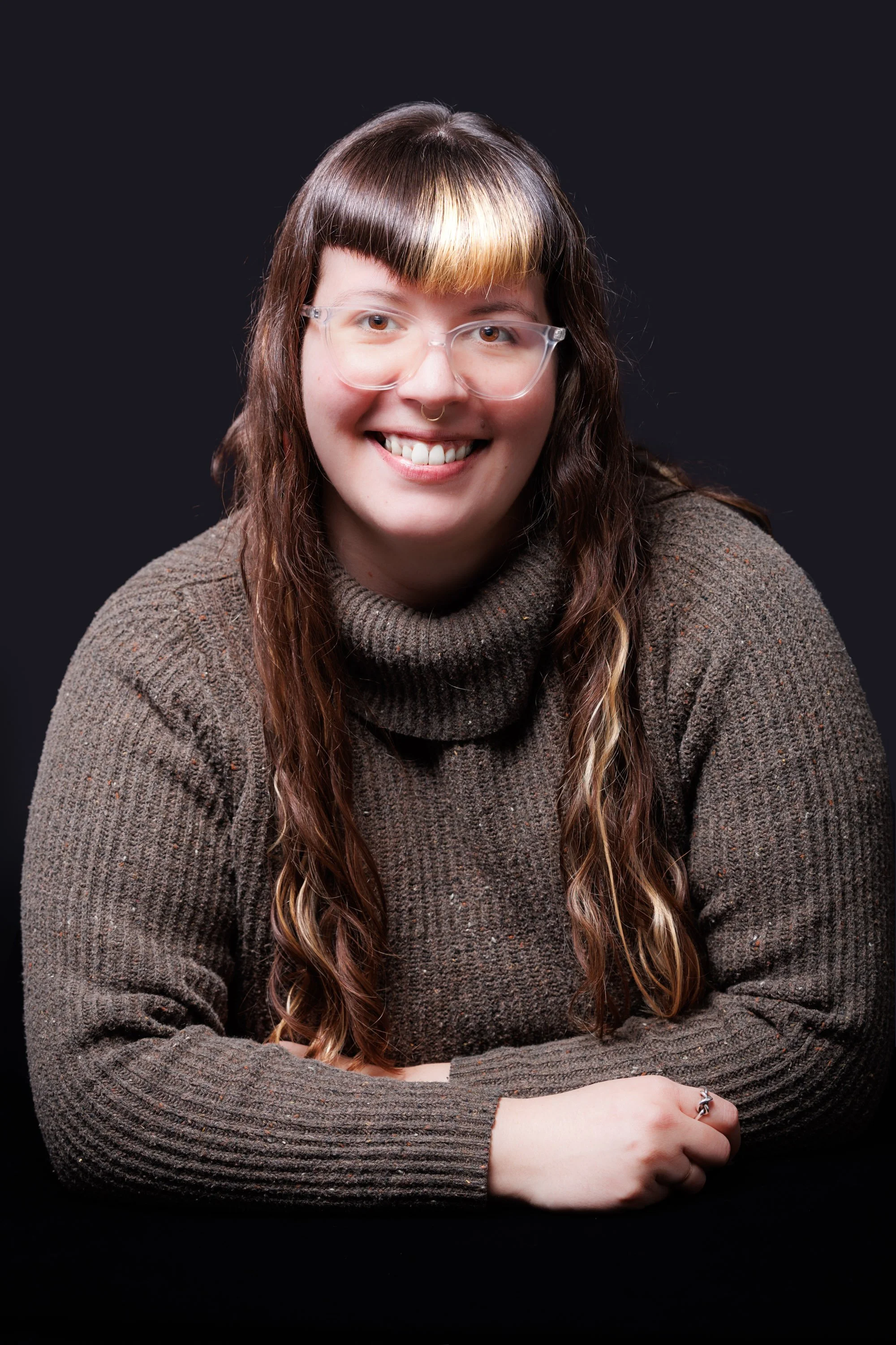 A woman with long wavy hair, wearing glasses and a brown turtleneck sweater, smiling at the camera against a dark background.
