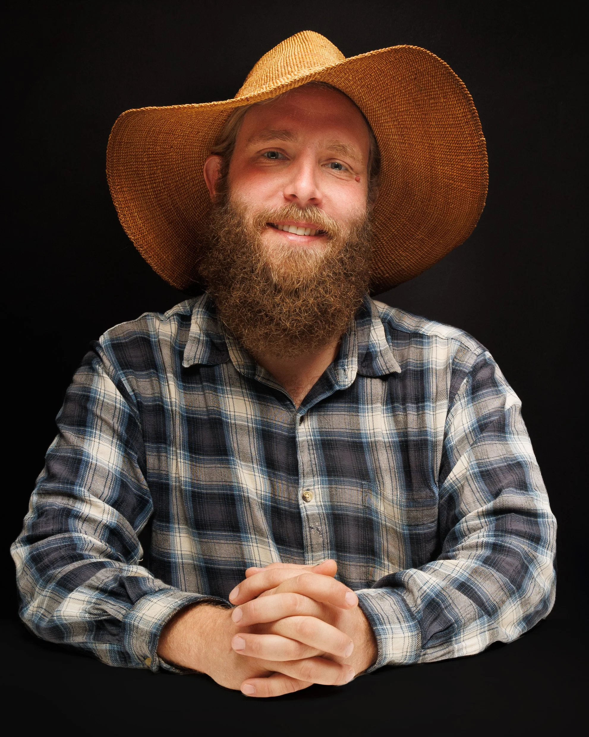 A man with a beard wearing a large straw hat and a plaid shirt, sitting against a black background.