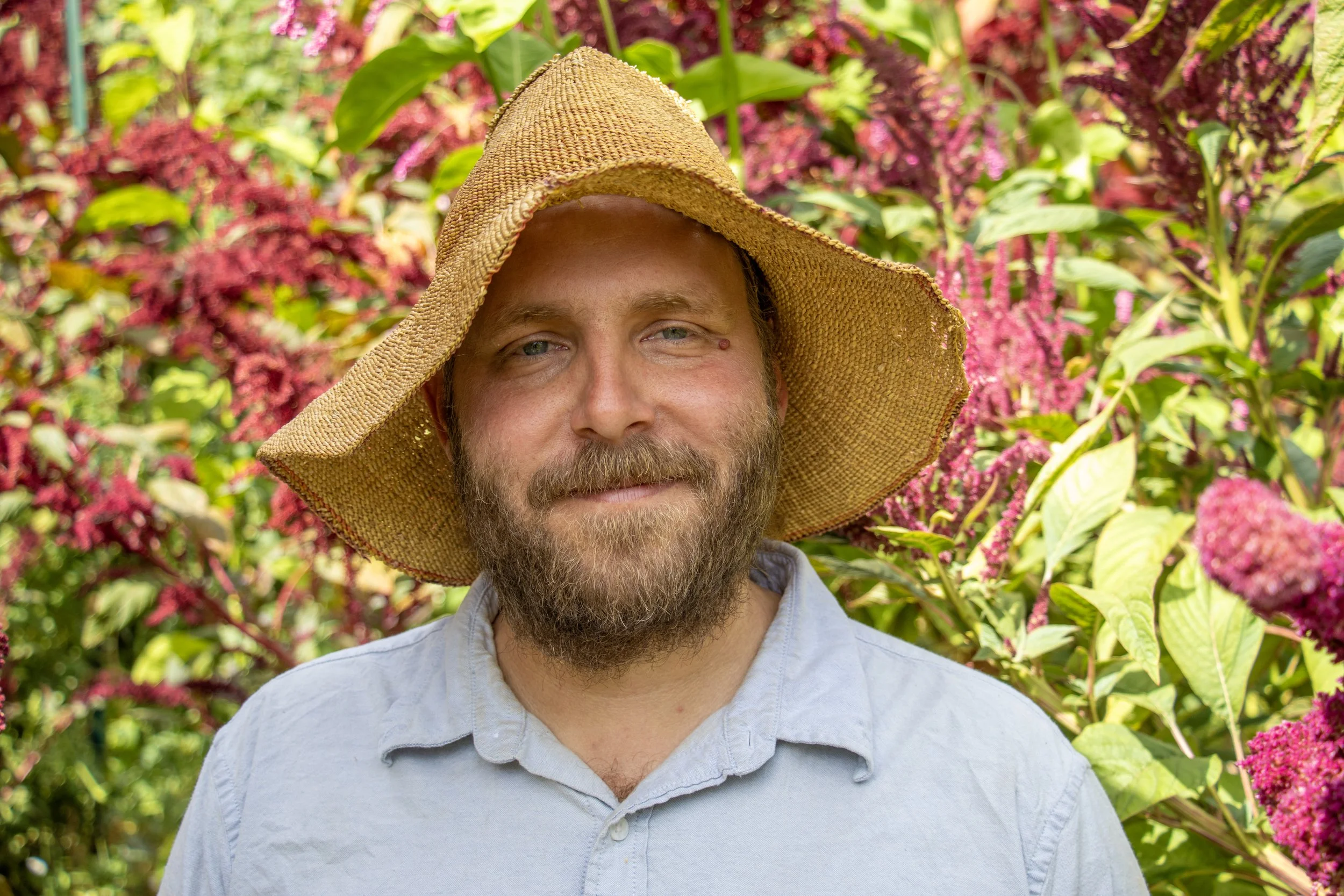 A man with a beard wearing a large straw hat and a plaid shirt, sitting against a black background.