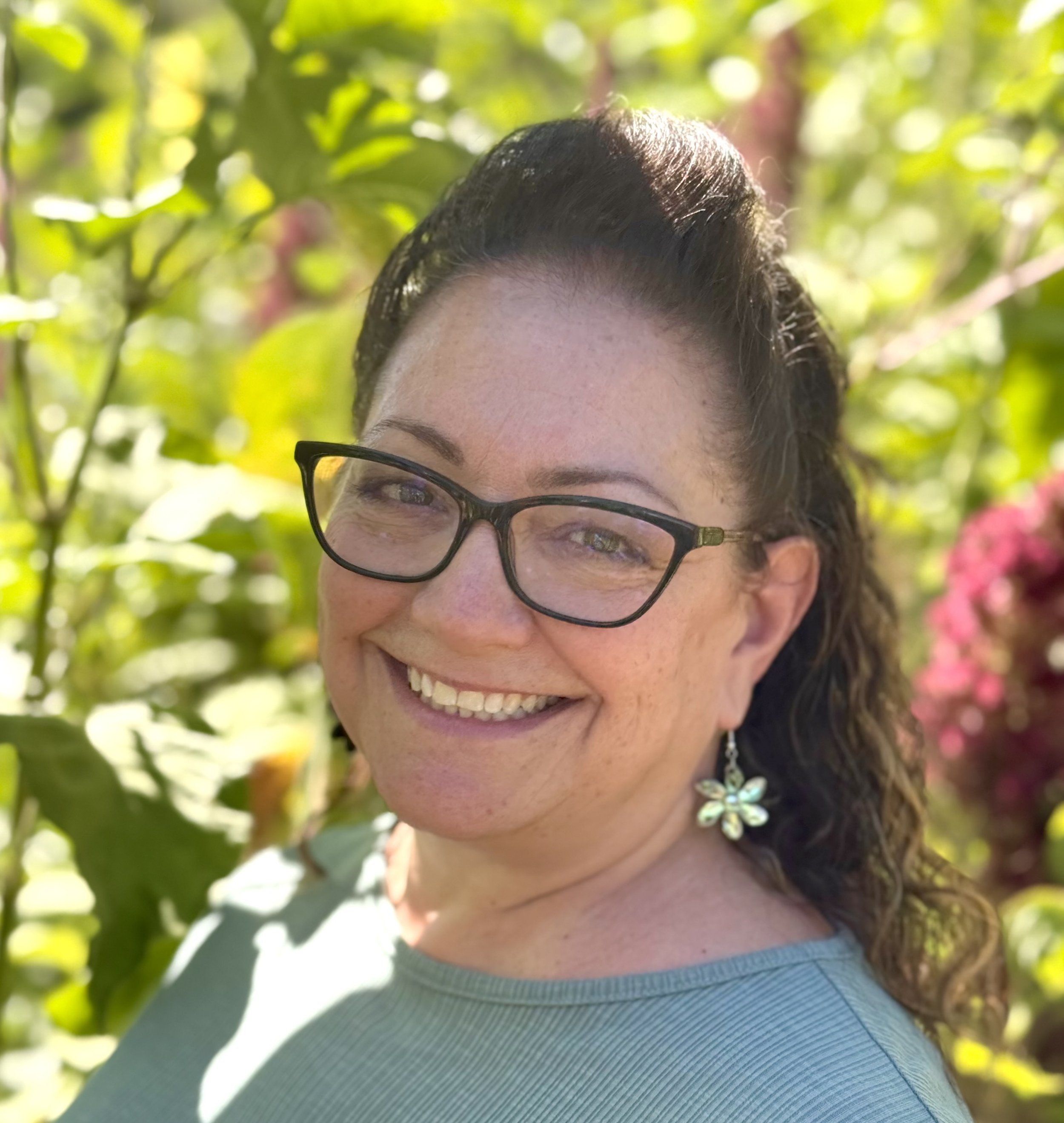 A woman with curly brown hair wearing glasses, a colorful floral patterned top, silver earrings, and smiling at the camera against a black background.