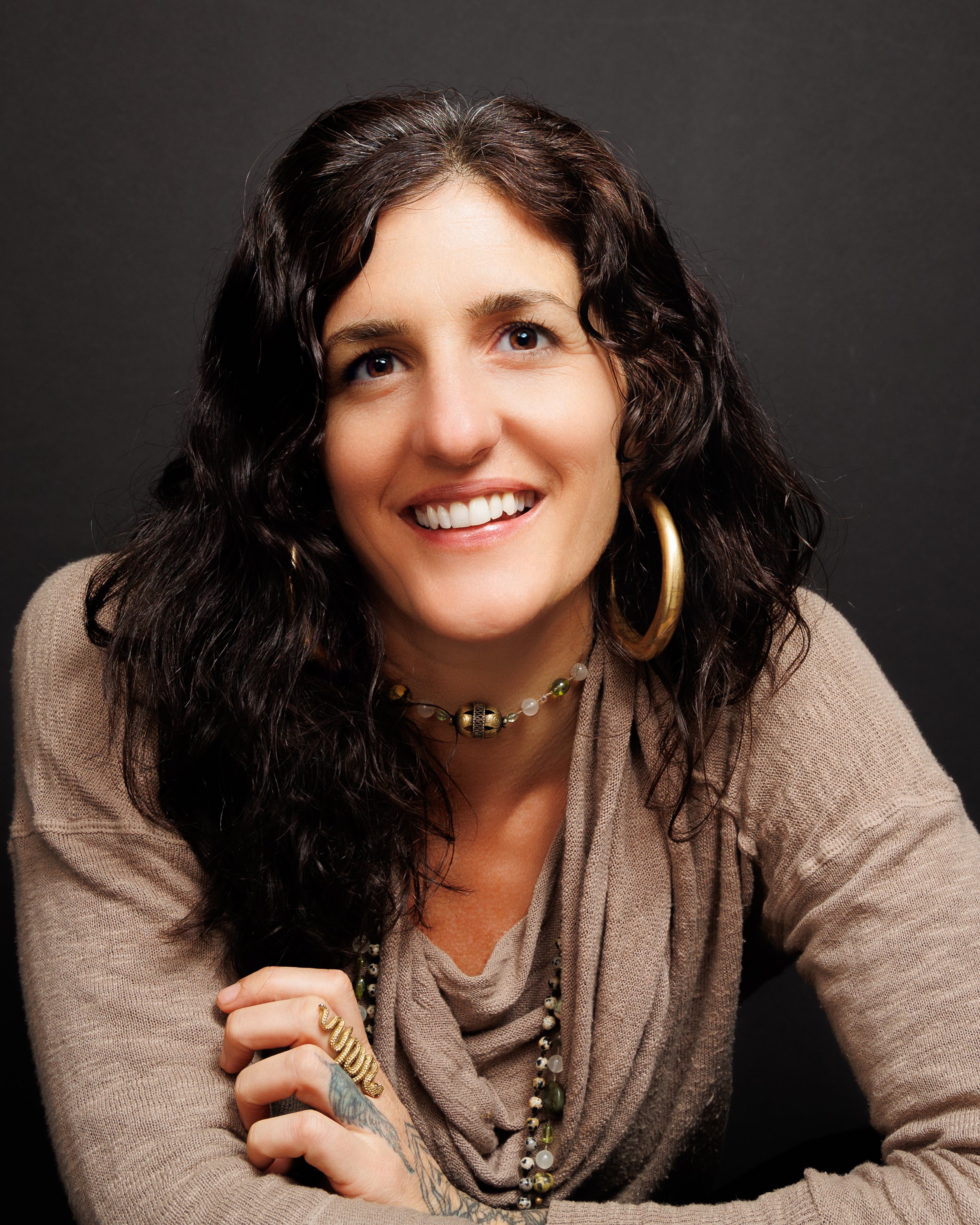 A woman with curly dark hair, wearing a beige top, large hoop earrings, and layered necklaces, smiling against a dark background.