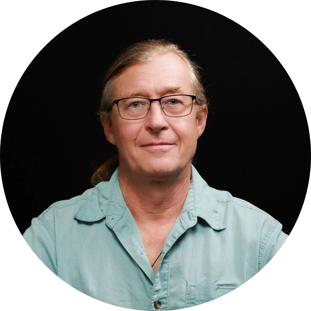 A middle-aged man with glasses, long hair tied back, wearing a light green shirt, smiling against a black background.
