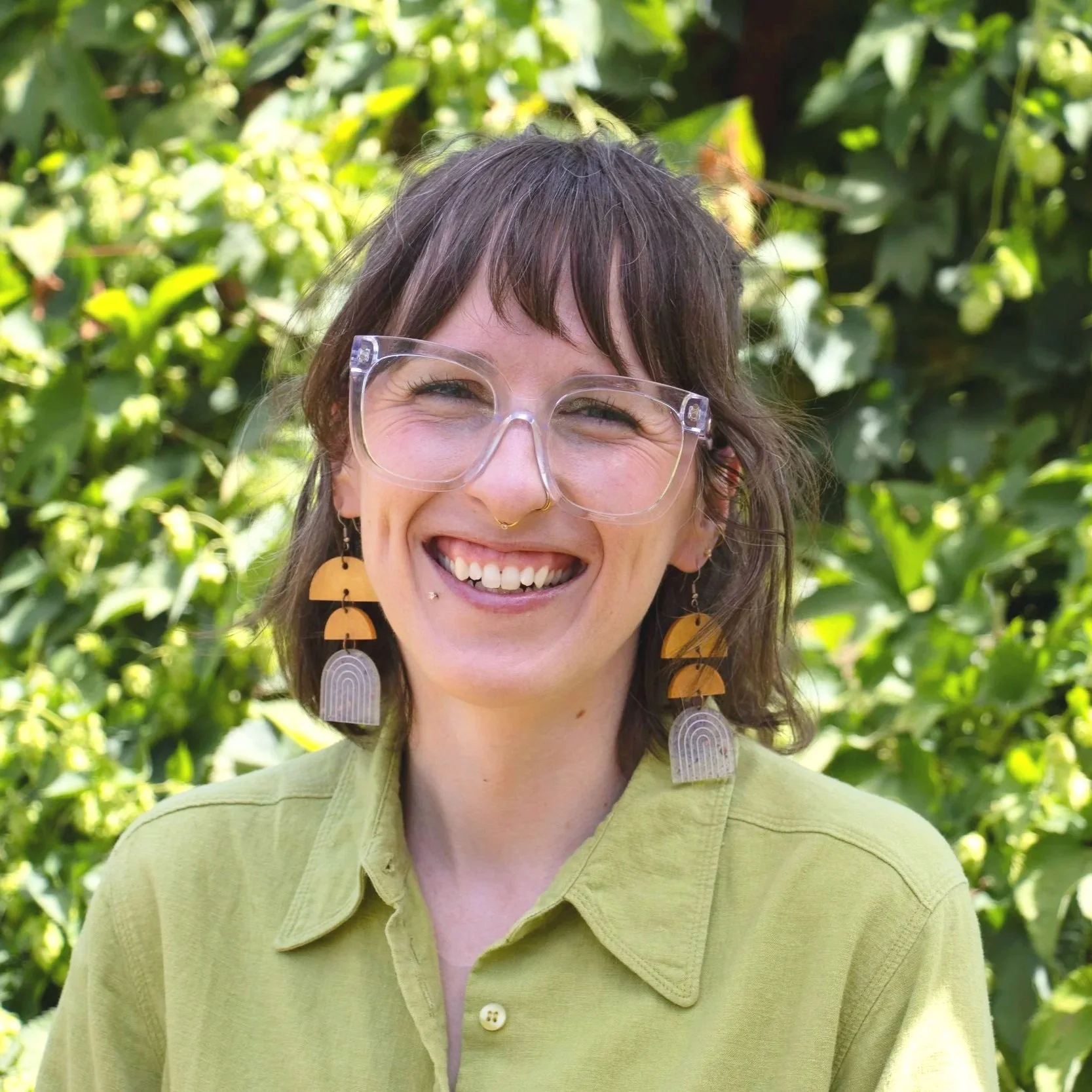 Portrait of a young woman with wavy brown hair, wearing glasses, a black blazer over a green top, and a distinctive necklace, sitting against a dark background.