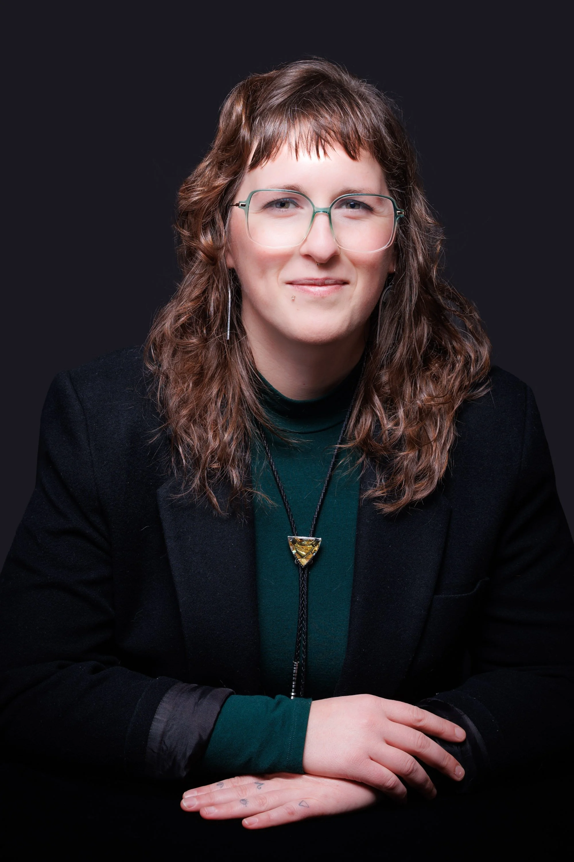 Portrait of a young woman with wavy brown hair, wearing glasses, a black blazer over a green top, and a distinctive necklace, sitting against a dark background.