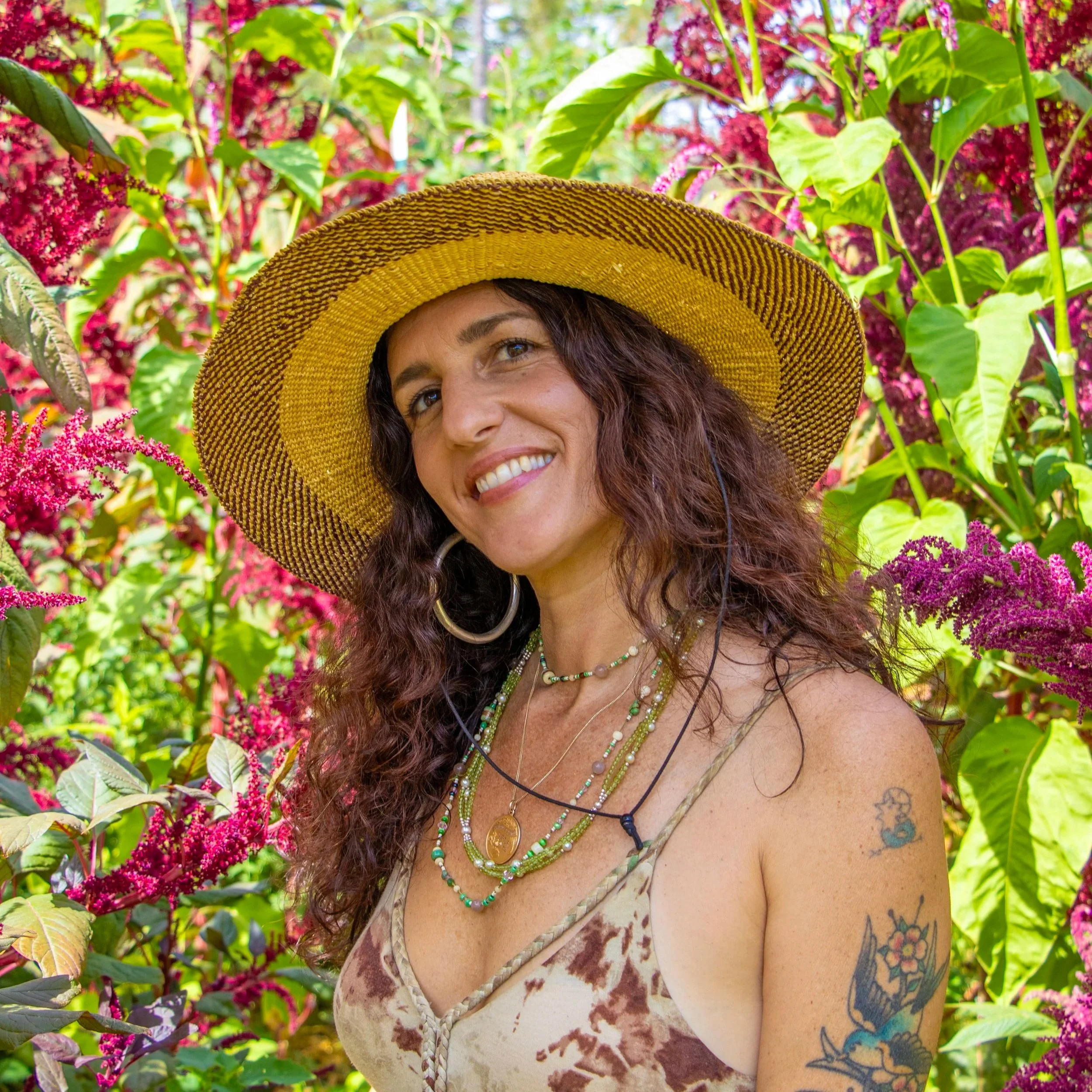 A woman with curly dark hair, wearing a beige top, large hoop earrings, and layered necklaces, smiling against a dark background.