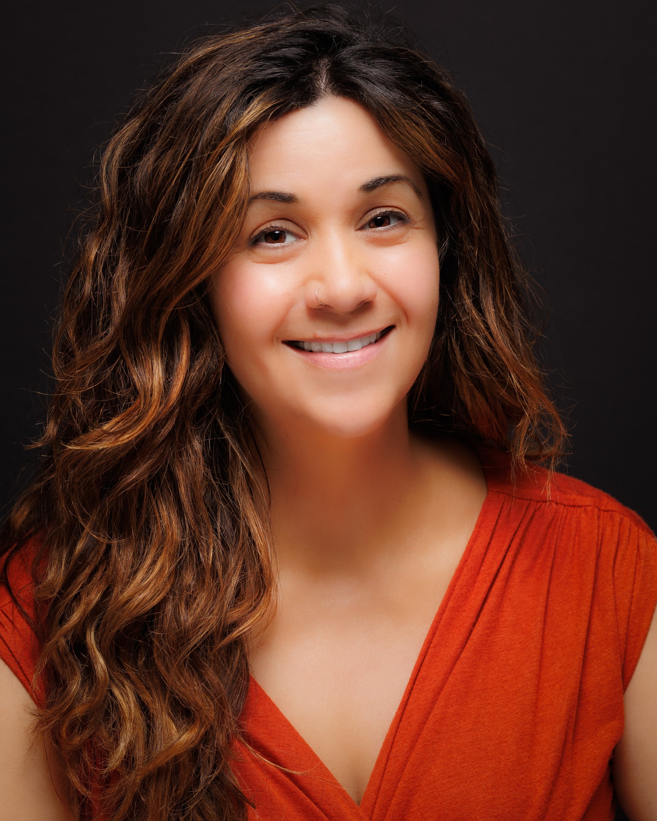 A woman with long wavy brown hair and a small nose ring is smiling and wearing an orange top against a black background.