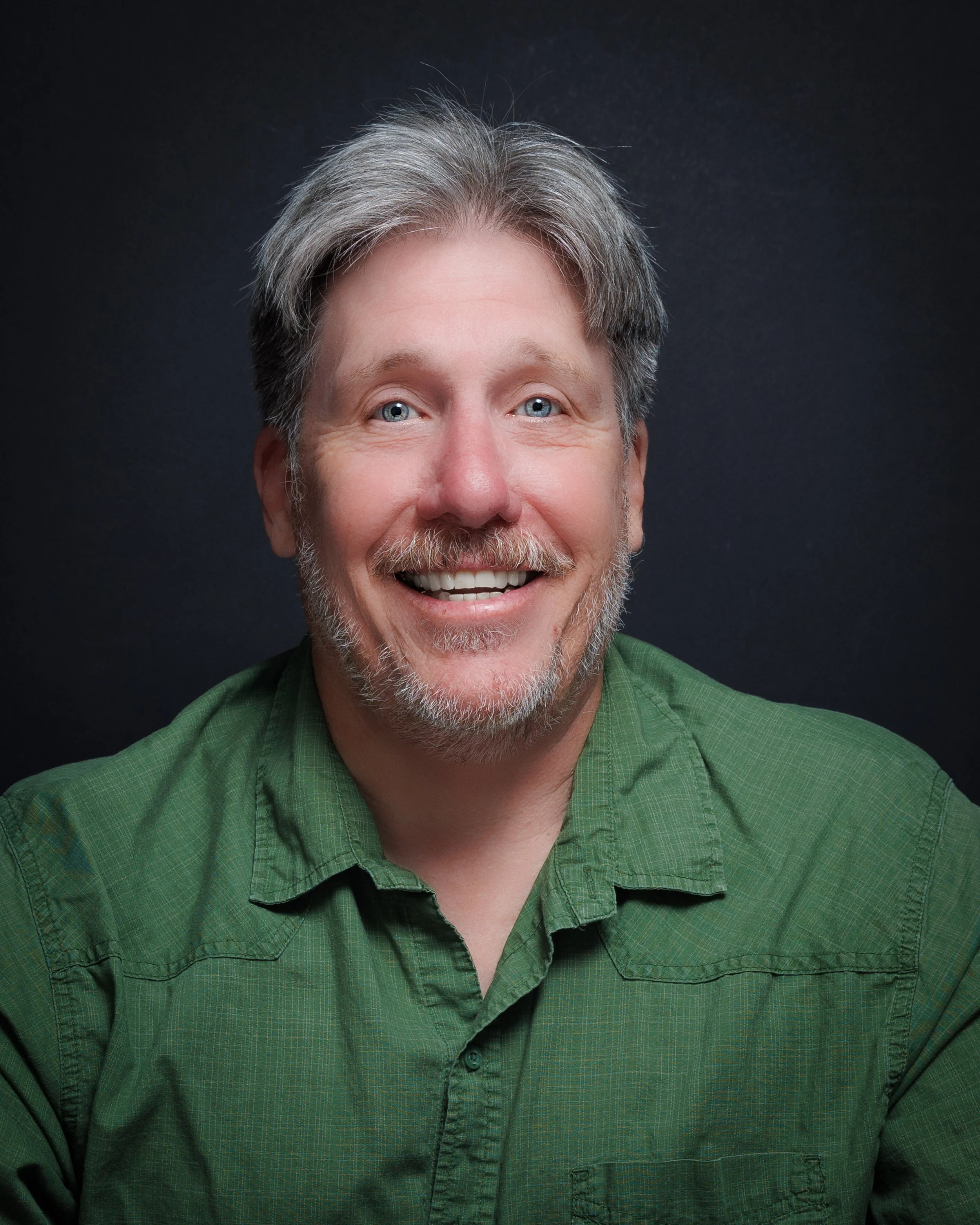 Headshot of a smiling middle-aged man with blue eyes, gray and brown hair, a beard, wearing a green button-up shirt against a dark background.
