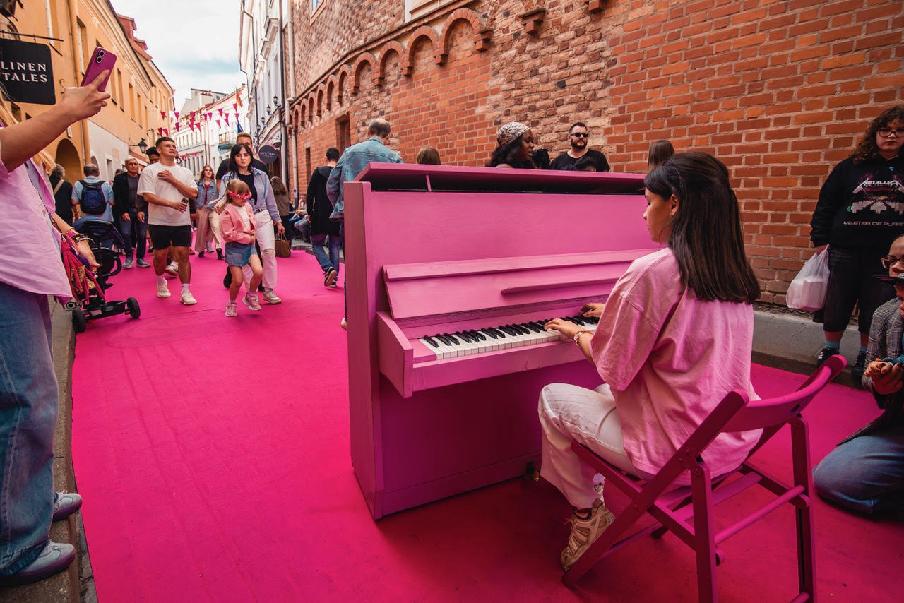 A woman playing a pink upright piano on a pink carpet in a busy outdoor street with people walking and taking photos, lined with brick walls and colorful buildings.