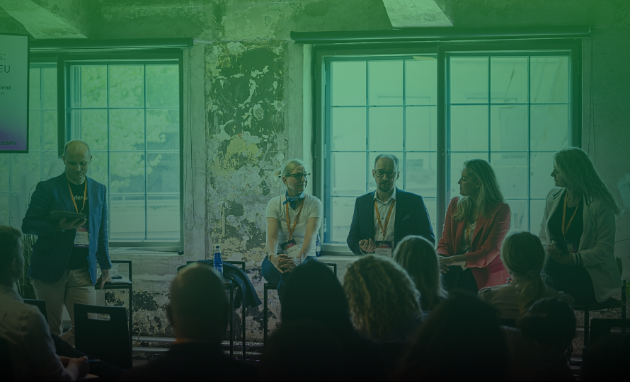 Panel of five people at a conference or seminar, speaking to an audience in a room with large windows and peeling wall paint.