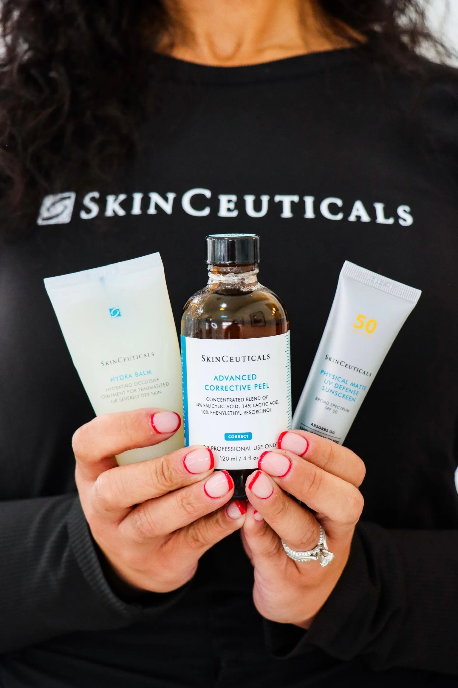 Person holding three skincare products from SkinCeuticals, with a black shirt featuring the brand's logo. The person has red and white nail polish and multiple rings on their fingers.