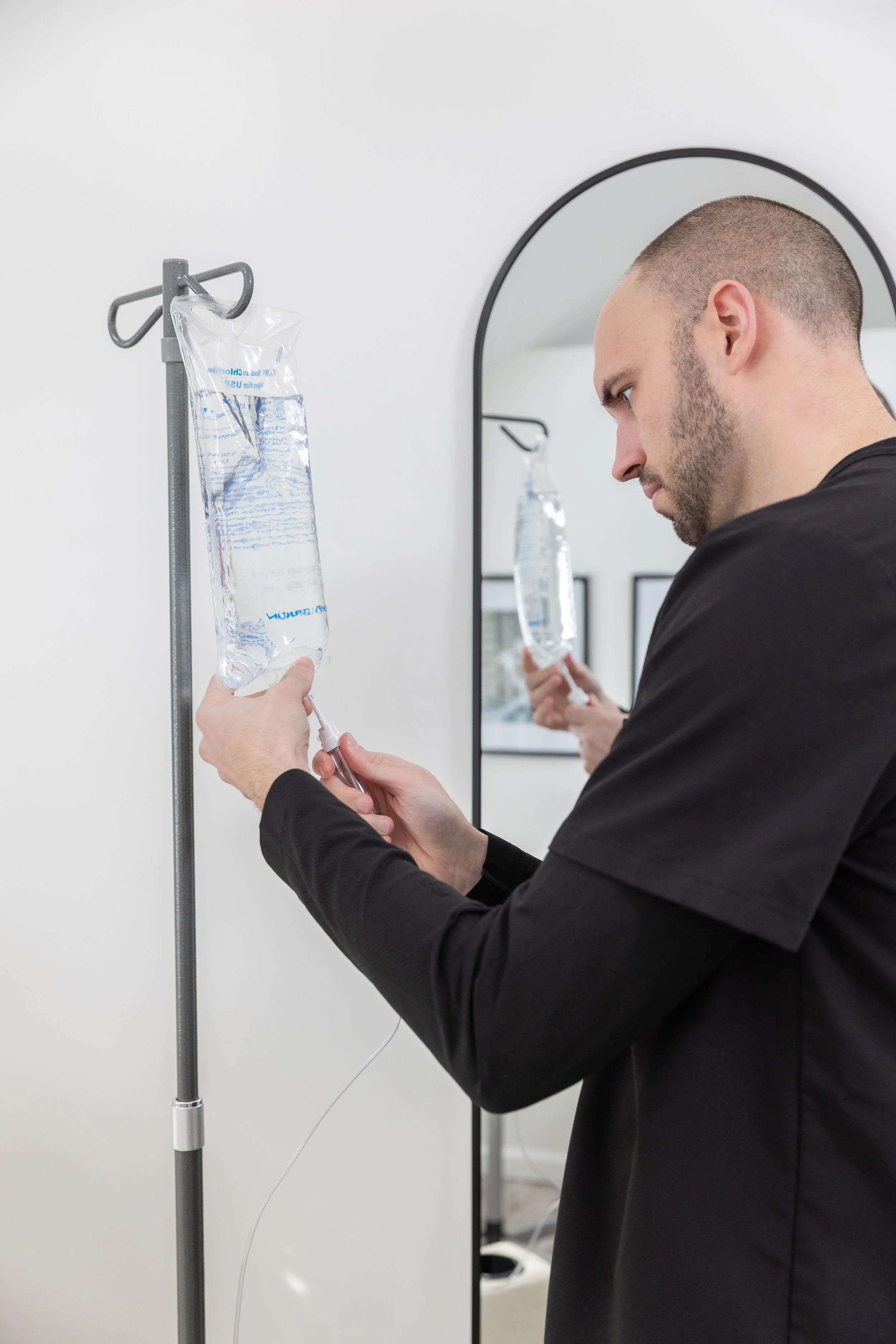 A man in a black shirt prepares an IV bag of saline solution for medical use, with a mirror and hospital setting in the background.