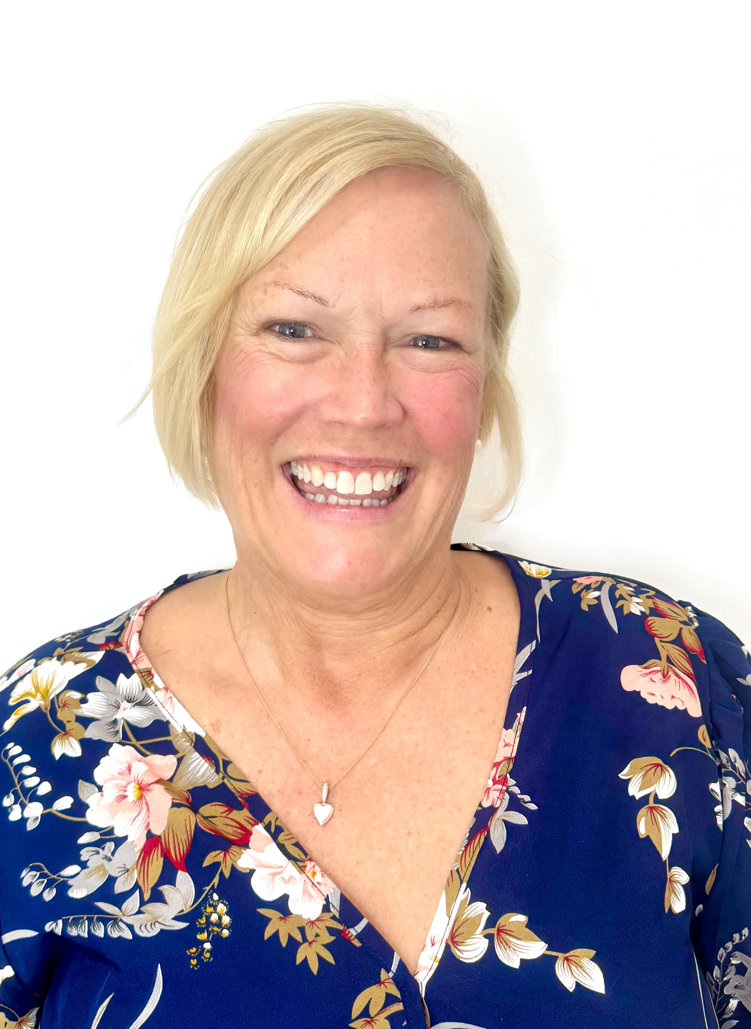 Close-up photograph of a smiling blonde woman with blue eyes, wearing a navy floral dress and a silver heart-shaped necklace, against a white background.