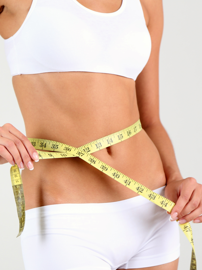 A woman wearing a white sports bra and white underwear measuring her waist with a yellow measuring tape.