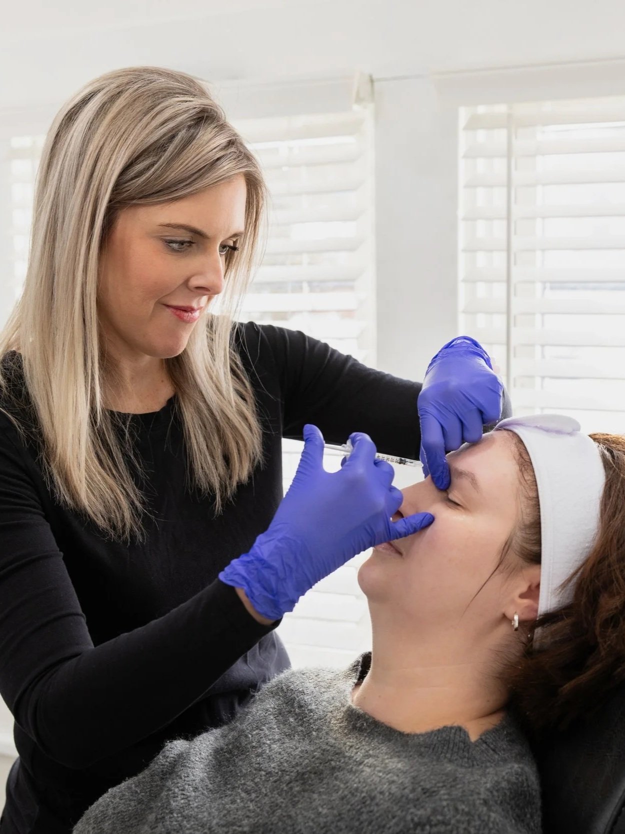 A woman is administering a cosmetic injection to another woman's forehead in a clinical setting. The healthcare worker is wearing blue gloves and a black shirt, and the patient has a headband and a gray sweater.