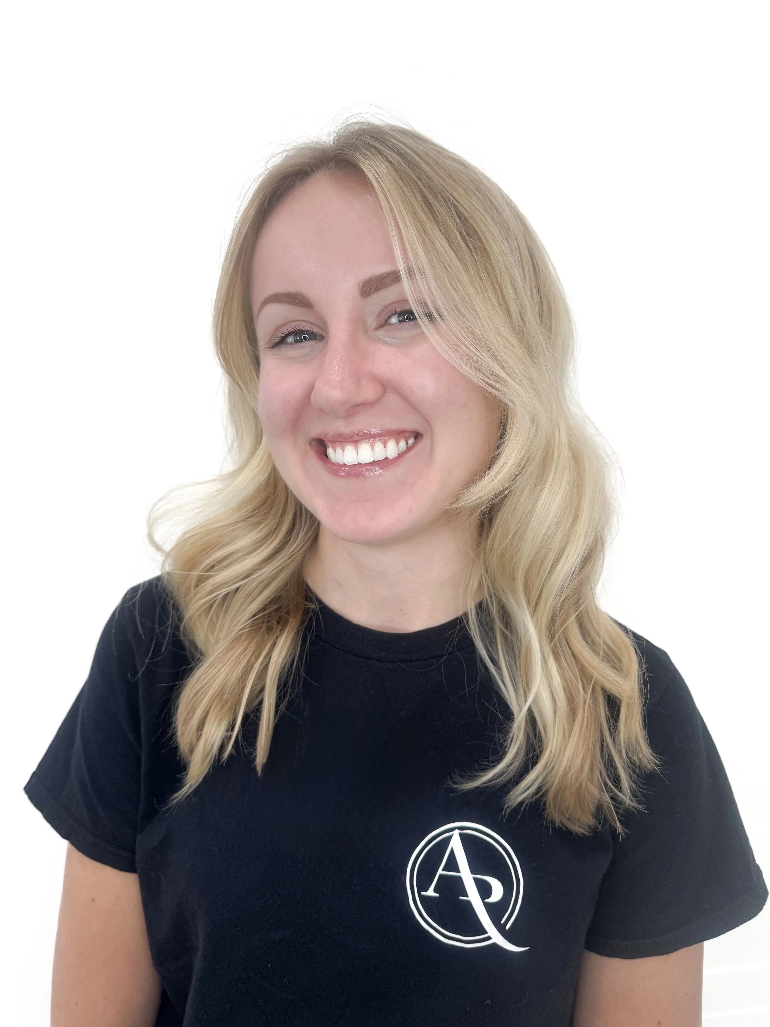 Close-up of a woman with blonde hair, smiling, wearing a black shirt with a white circular logo featuring the Greek letters Alpha and Phi.