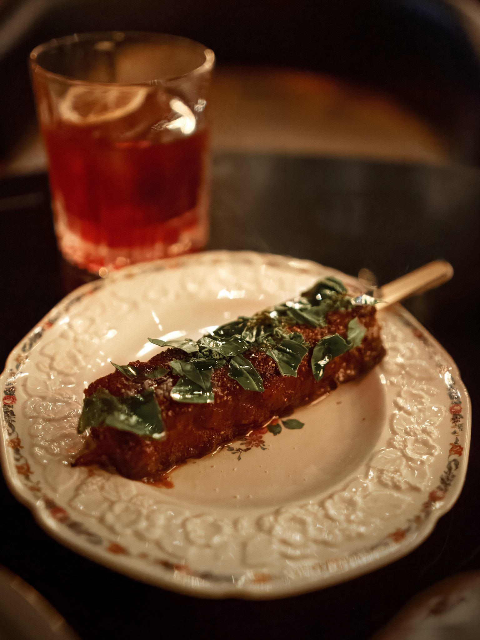 Pork belly skewer garnished with green herbs on a decorative white vintage plate, with a glass of red cocktail in the background.