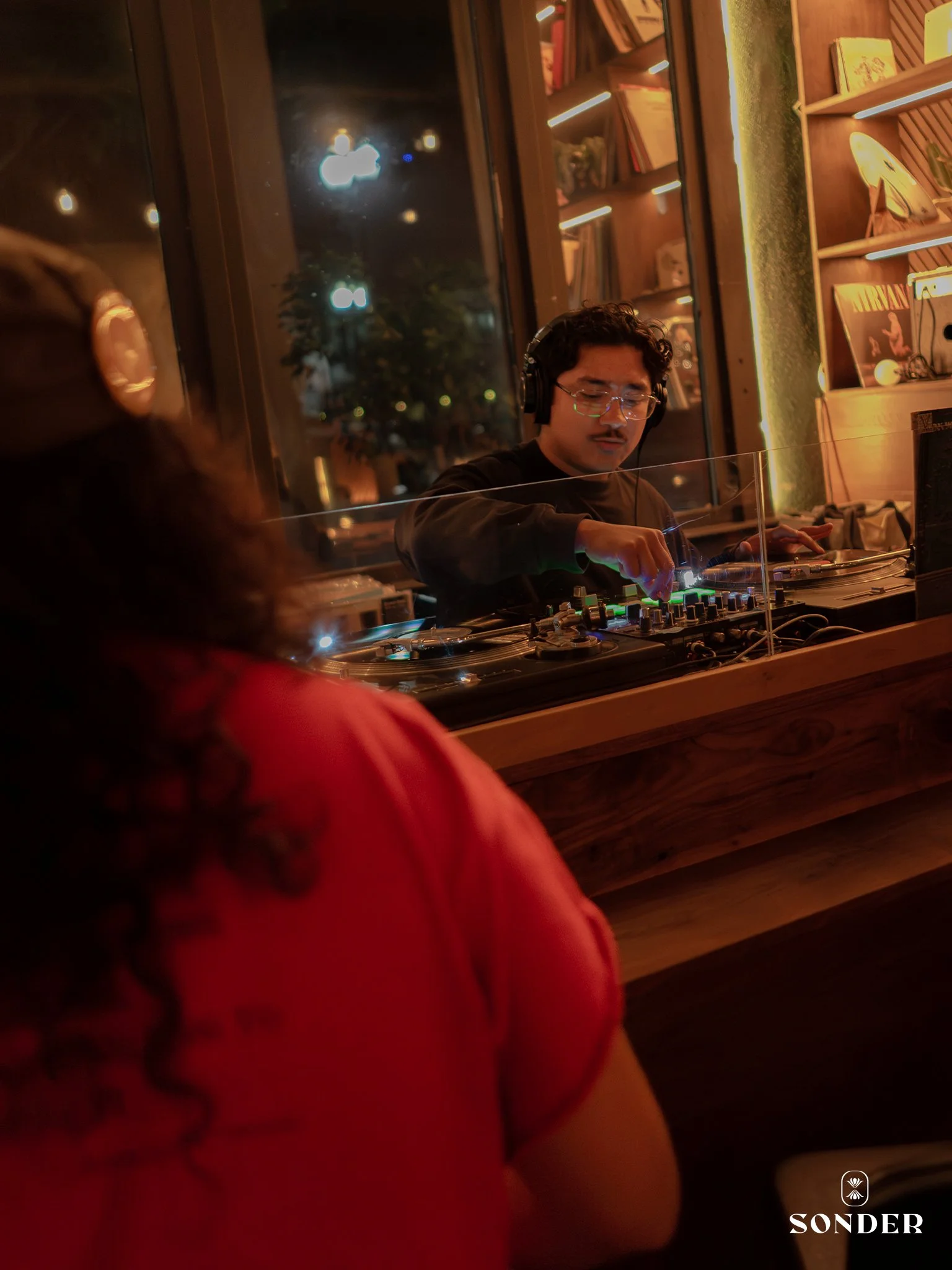 A DJ with curly hair, glasses, and headphones performing at a bar or club at night, with shelves of books and vinyl records in the background. A guest with dark, curly hair wearing a red top is in the foreground.