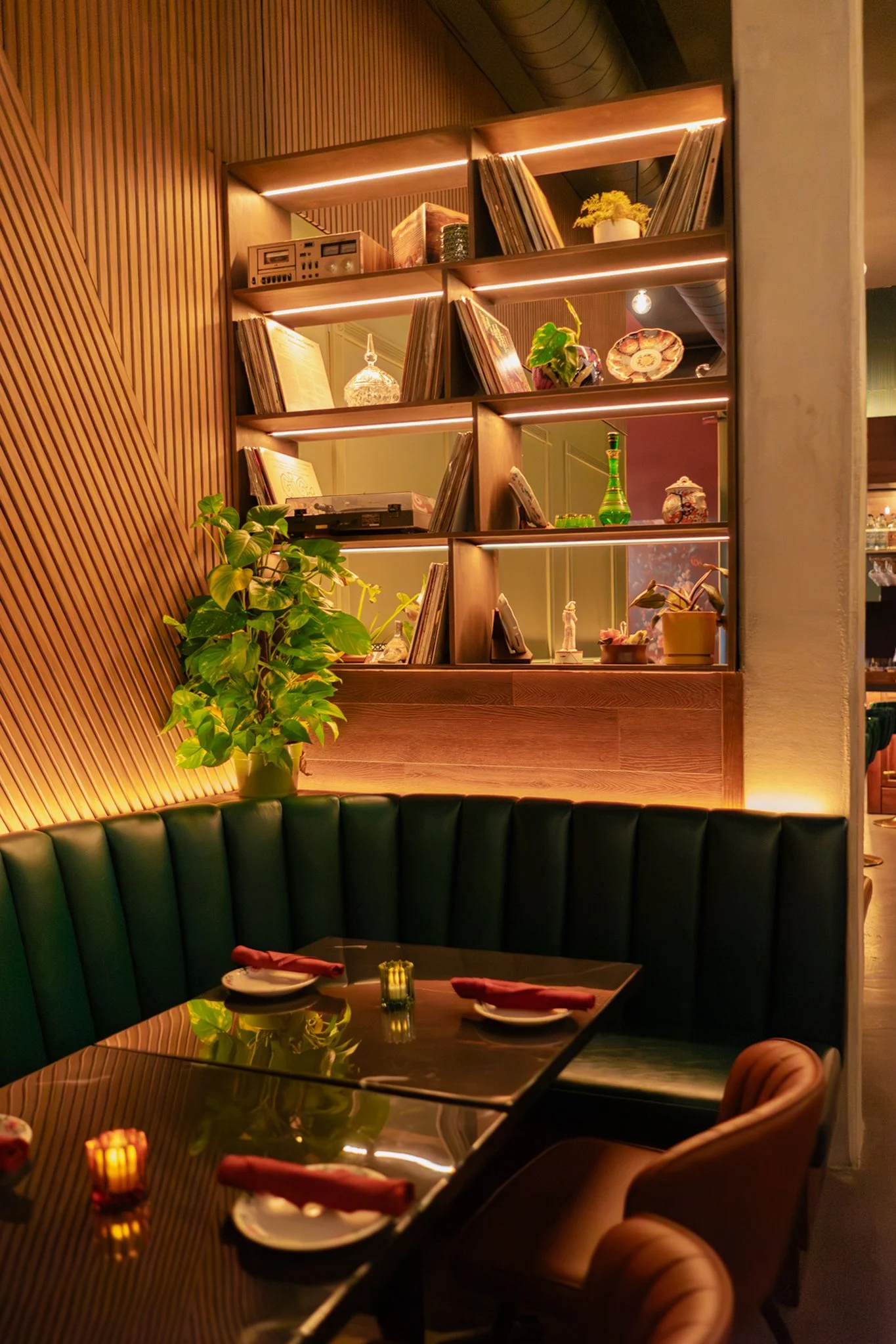A cozy restaurant corner with a dark green leather booth, a wooden table set for four with plates, red napkins, and candle holders, and decorative plants on shelves against a wood-paneled wall.