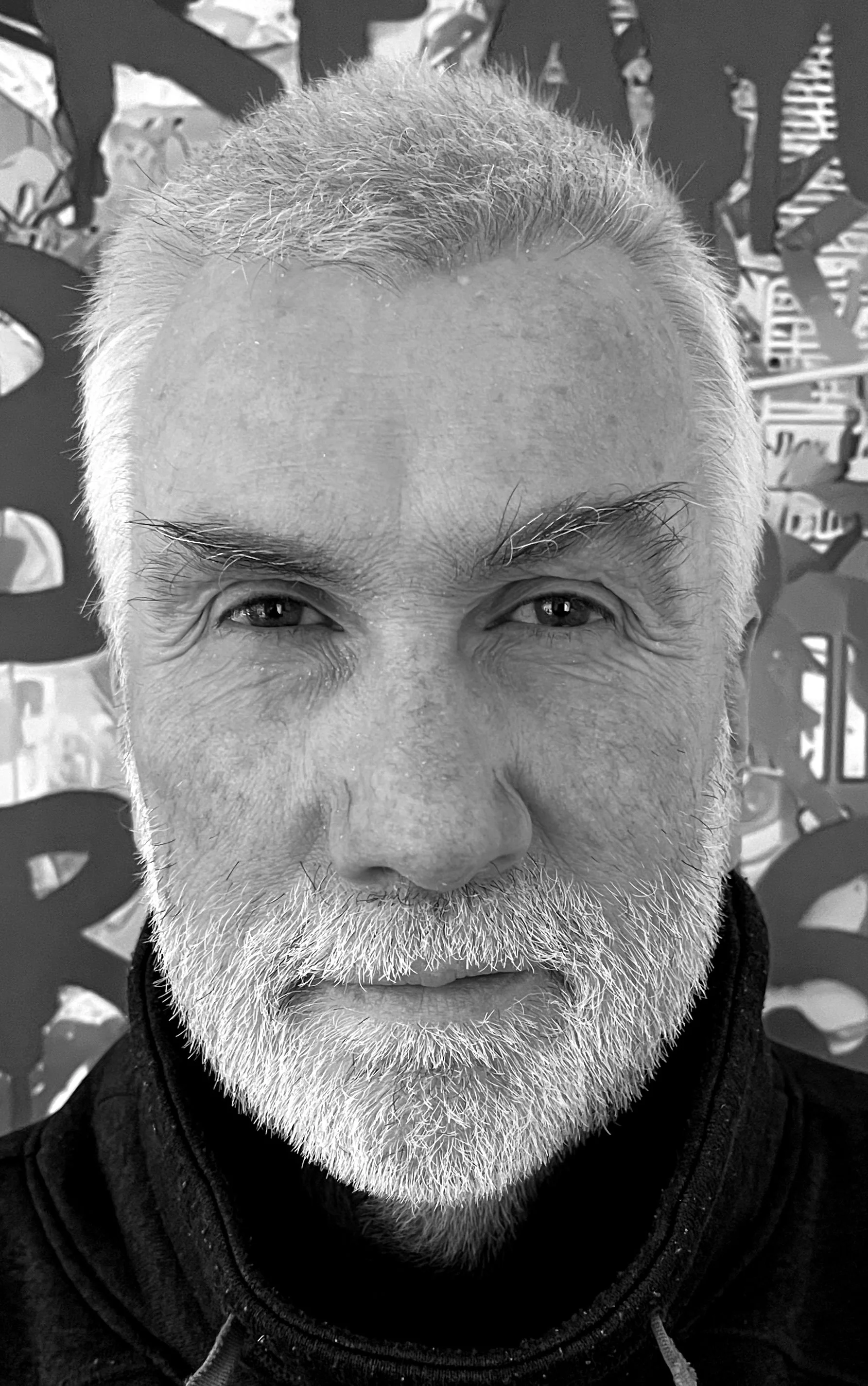 Close-up black and white photo of an older man with a full white beard and mustache, smiling slightly, with a patterned background.