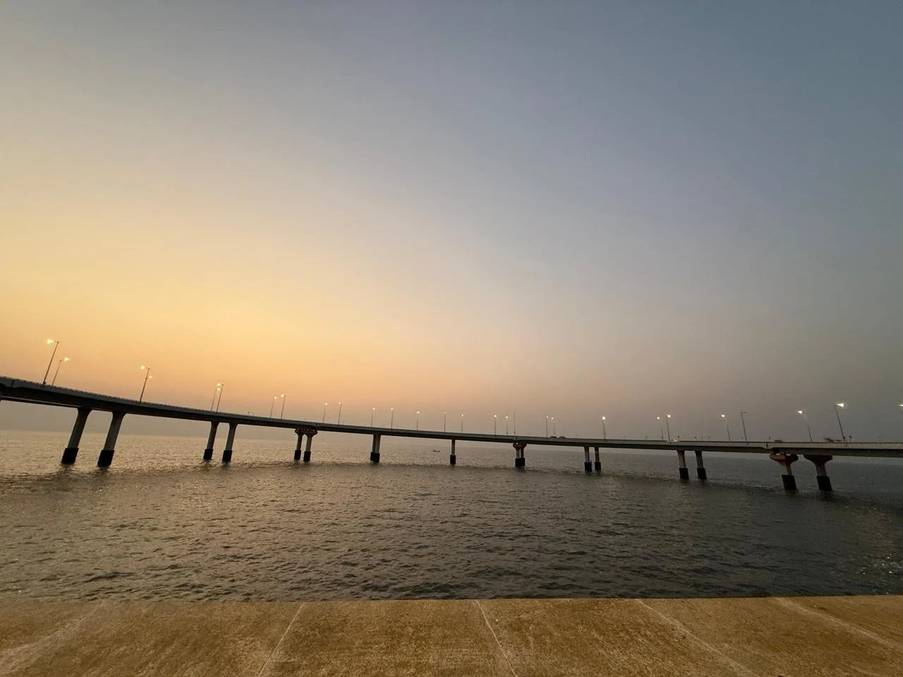 A long bridge extending over water during sunset with streetlights illuminated and a wooden or stone platform in the foreground.