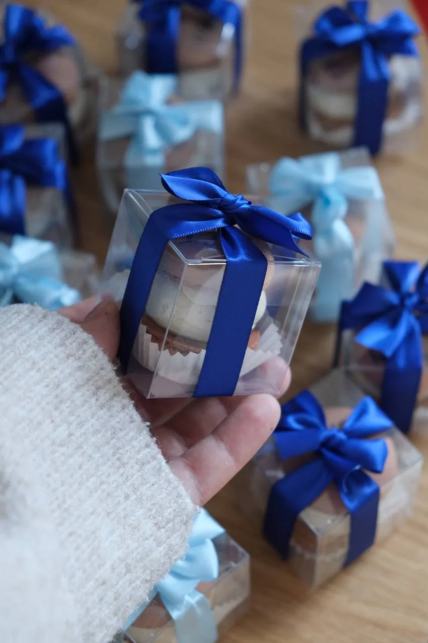 A hand holding a single boxed macaron tied with a royal-blue ribbon, more boxes on the desk