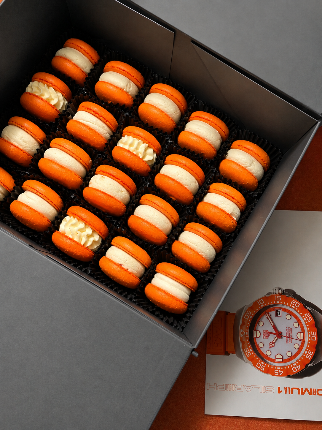 A box of orange macarons with white filling, some filled with cream, placed in black dividers, and a watch colored orange and white beside a paper.