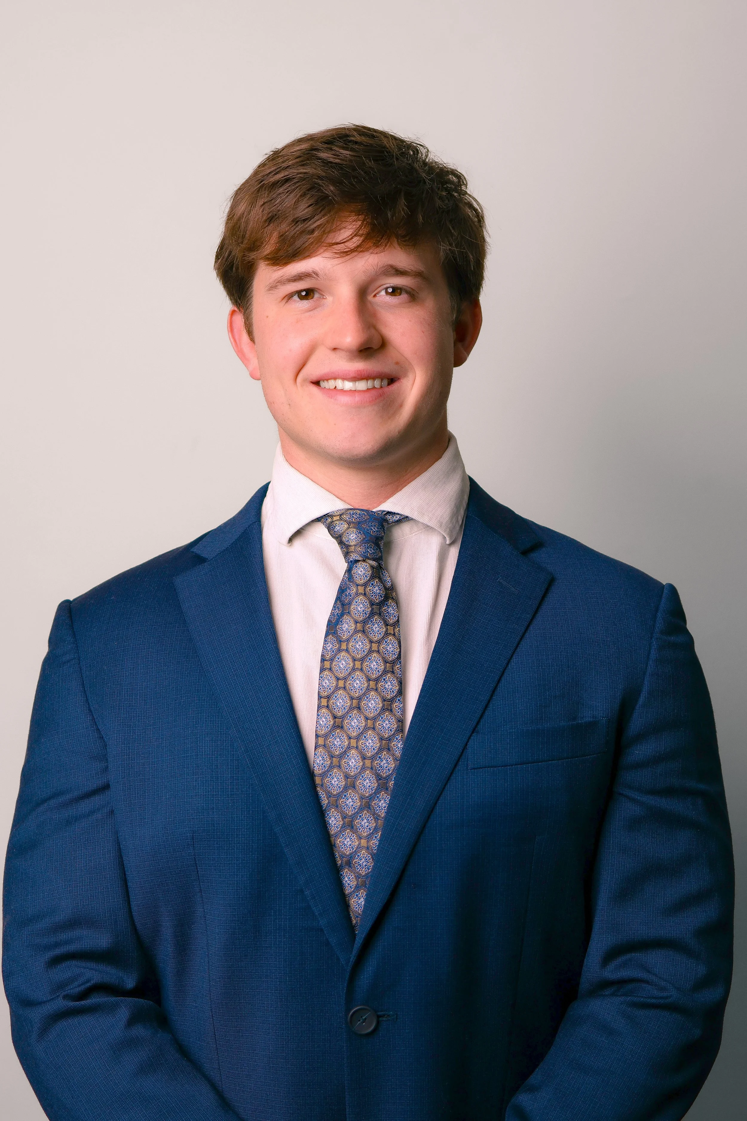 Young man with brown hair smiling, dressed in a black suit, white shirt, and red polka dot tie, against a gray background.