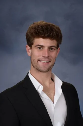 Young man with dark, curly hair wearing a green and white striped sweater and a silver necklace standing in front of a neon-lit artwork on a pink wall.