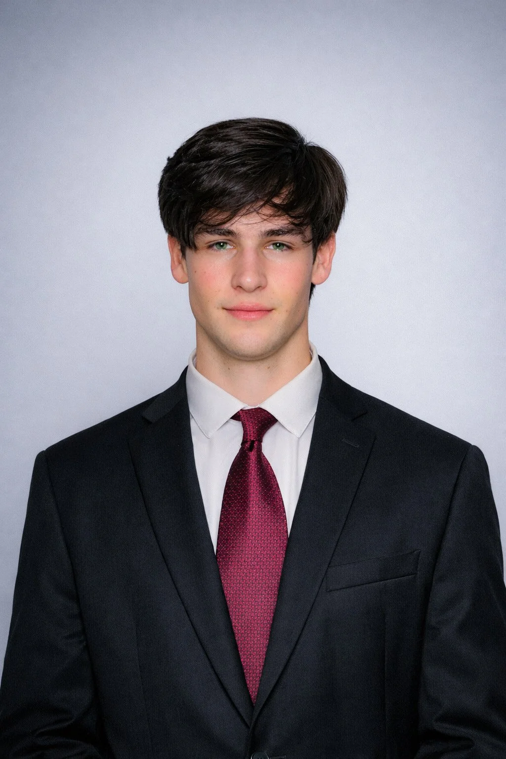 Young man wearing a black suit, white shirt, and red tie posed against a plain light gray background.