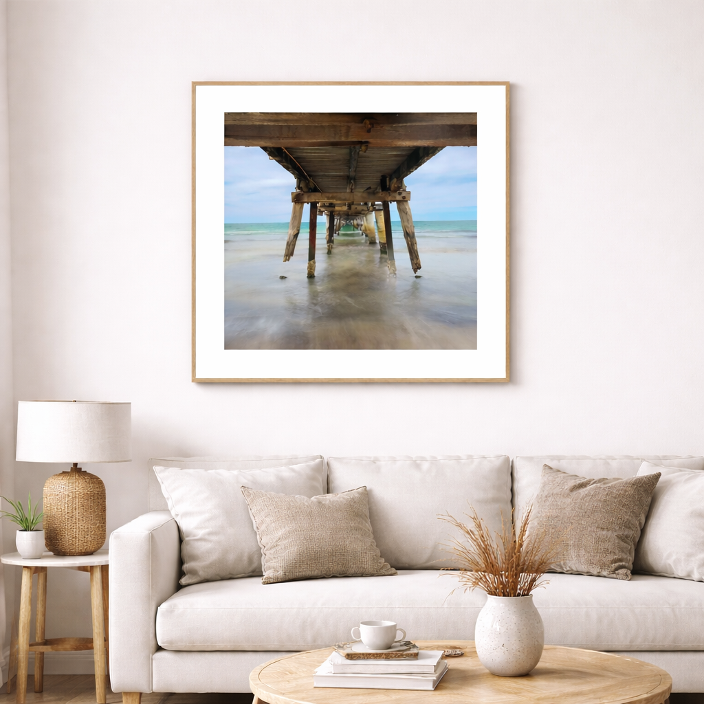 Square photographic print showing timber pylons beneath Tumby Bay Jetty at low tide, South Australia, displayed framed in a light, minimalist interior.