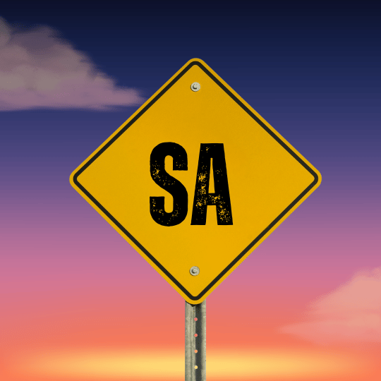 Yellow diamond-shaped road sign with black letters 'SA' against a colorful sky background at sunset.