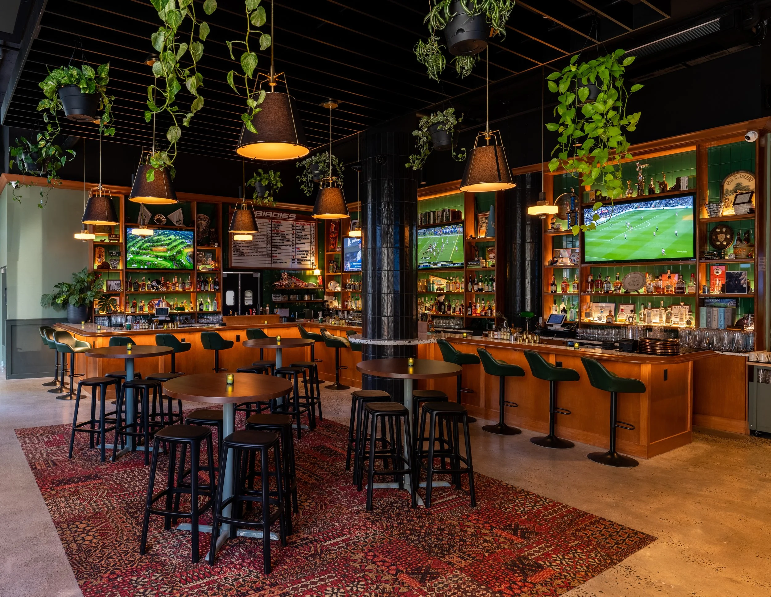 A modern bar with multiple television screens showing sports, wooden bar counter with barstools, small tables with chairs, hanging pendant lights, green plants hanging from ceiling, and a red patterned rug on polished concrete floor.