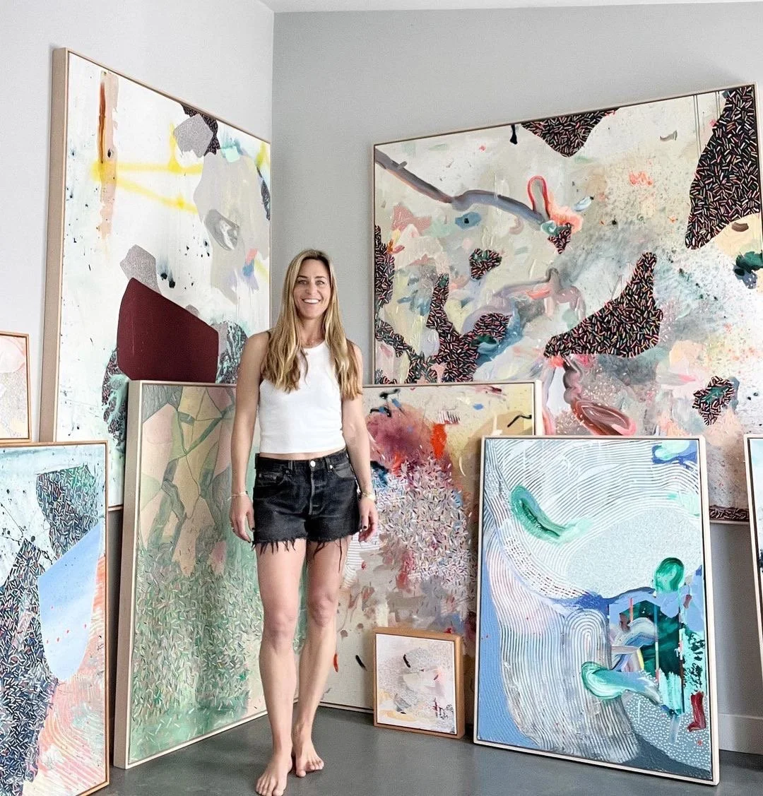 A woman standing barefoot in front of multiple abstract paintings leaning against a wall.