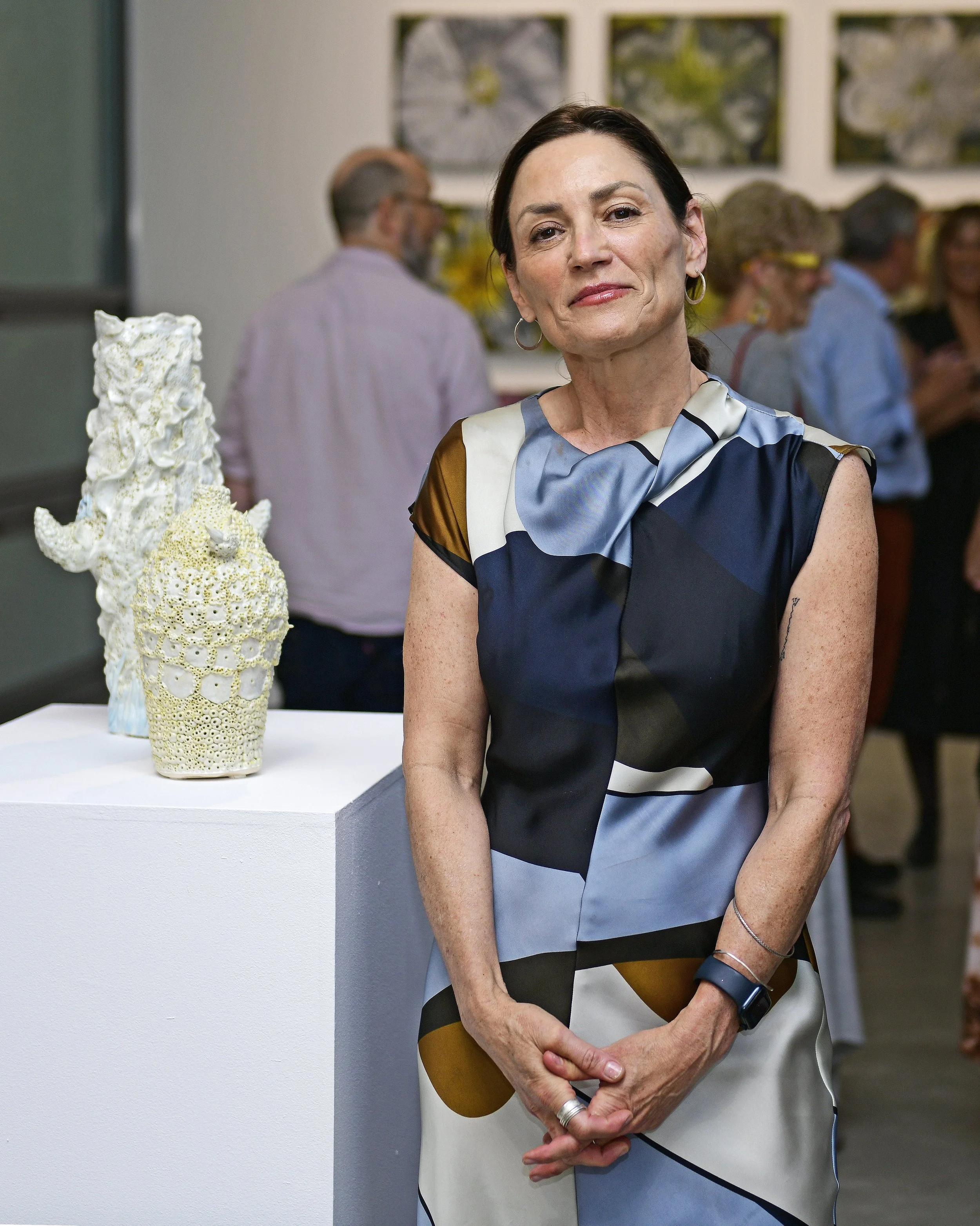 A smiling woman with dark hair and hoop earrings standing in front of a table with a textured sculpture. In the background, there are people socializing in an art gallery or exhibition setting.