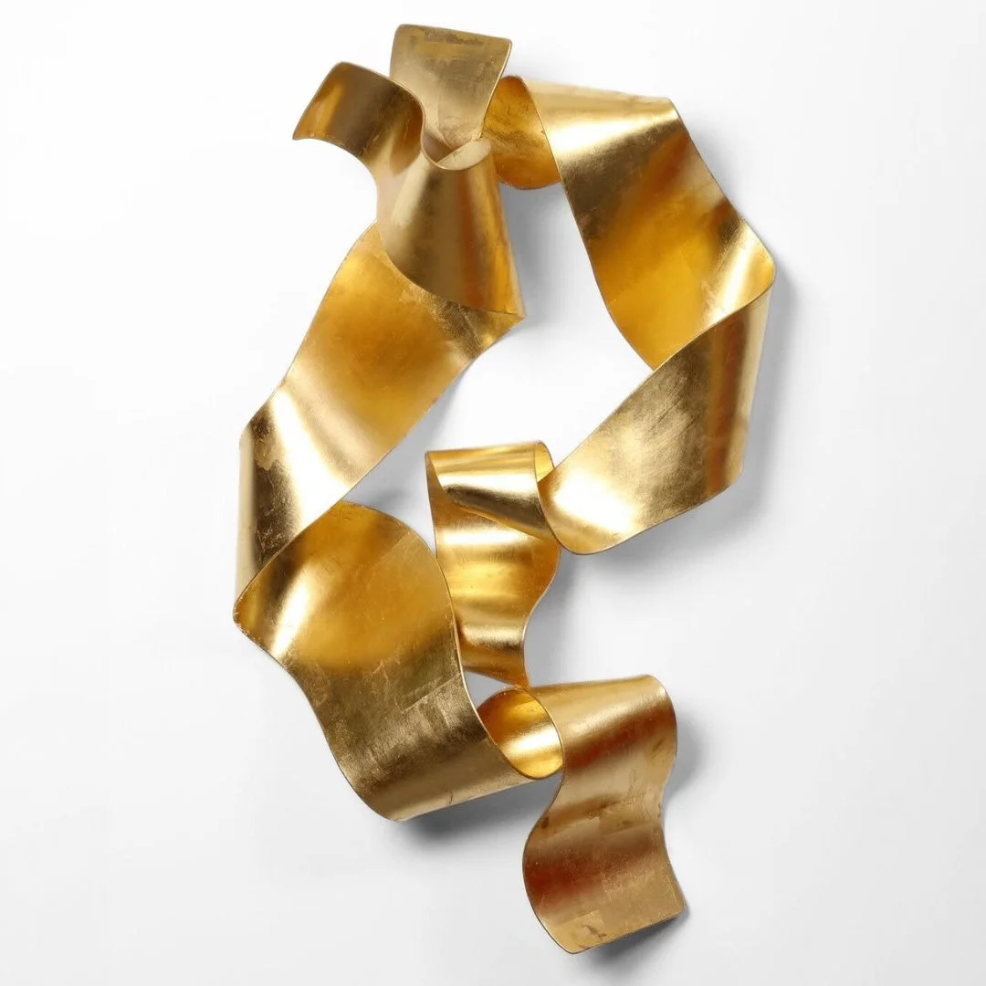A decorative gold metallic ribbon sculpture with wavy, curled shapes on a white background.