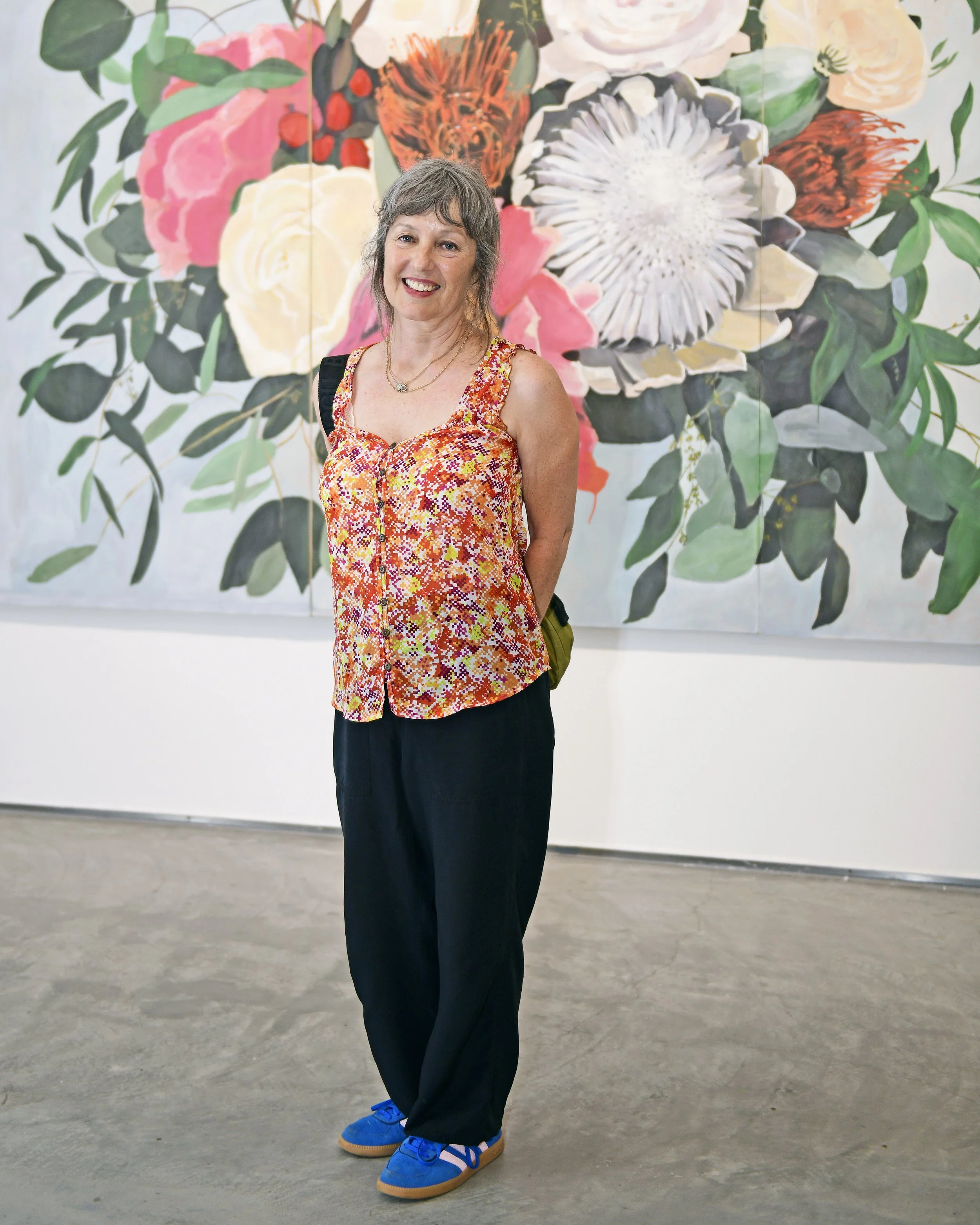 A smiling woman with short gray hair, wearing a sleeveless orange patterned top, black pants, and blue sneakers, standing in front of a large floral artwork with pink, white, orange, and green flowers and leaves.