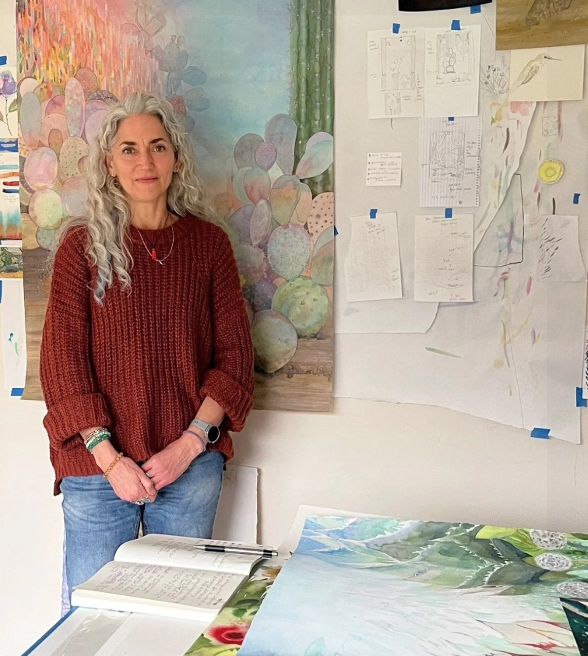 A woman with curly gray hair wearing a red knit sweater, standing in front of a wall with colorful artwork and sketches taped to it. She has her hands clasped and is standing next to a table with open notebooks and paintings.
