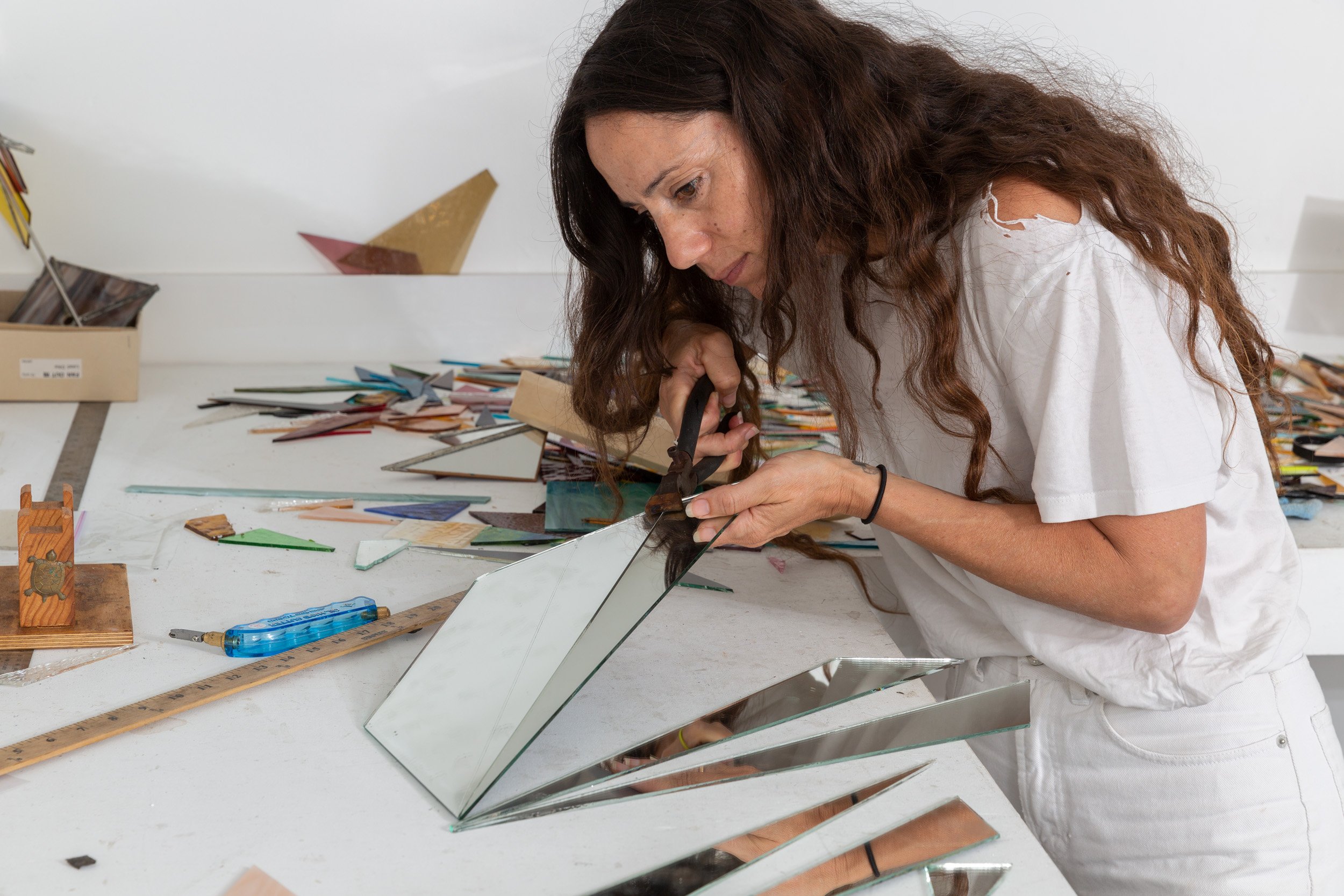 A woman with long curly dark hair working with glass and tools on a work table covered with various small glass pieces and crafting supplies in an art studio.