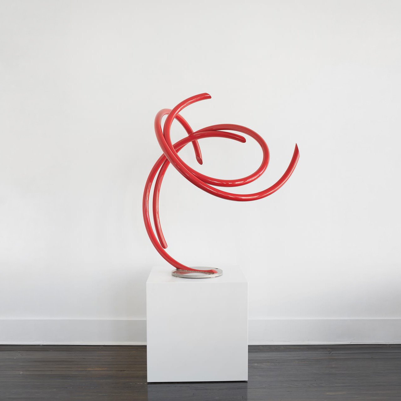 Red abstract sculpture with curving tubular forms on a white pedestal against a white wall.