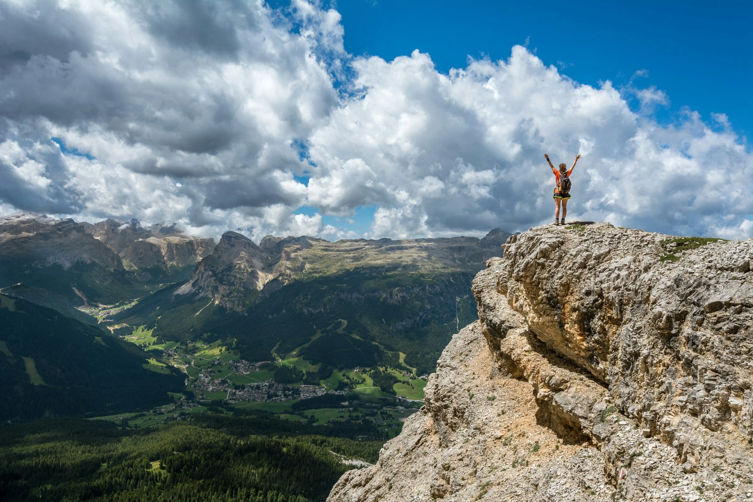 A person standing on a rocky ledge on a mountain, overlooking a valley with green fields and distant mountains under a partly cloudy sky.